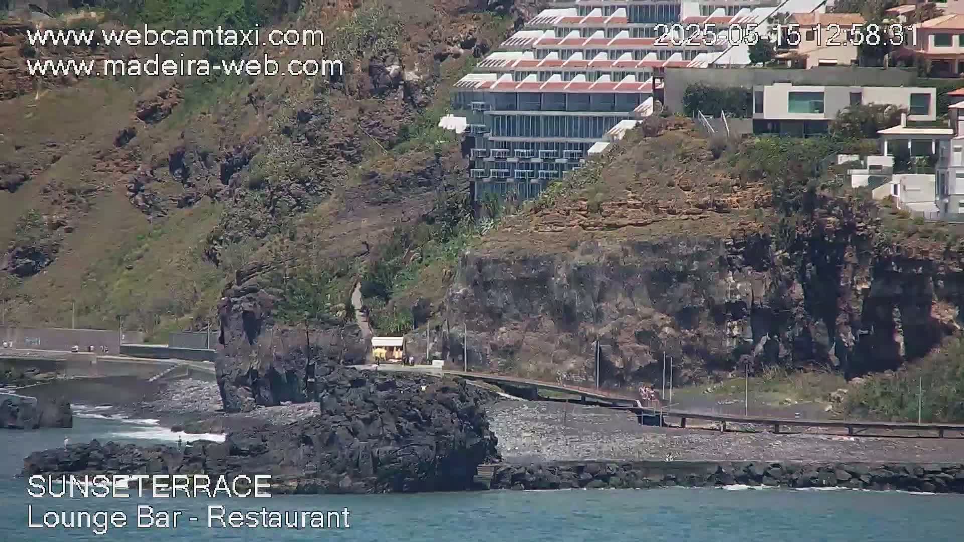 A rocky, dark-colored shore with a building perched on a cliff above, and calm ocean waves lapping at the base of the rocks.