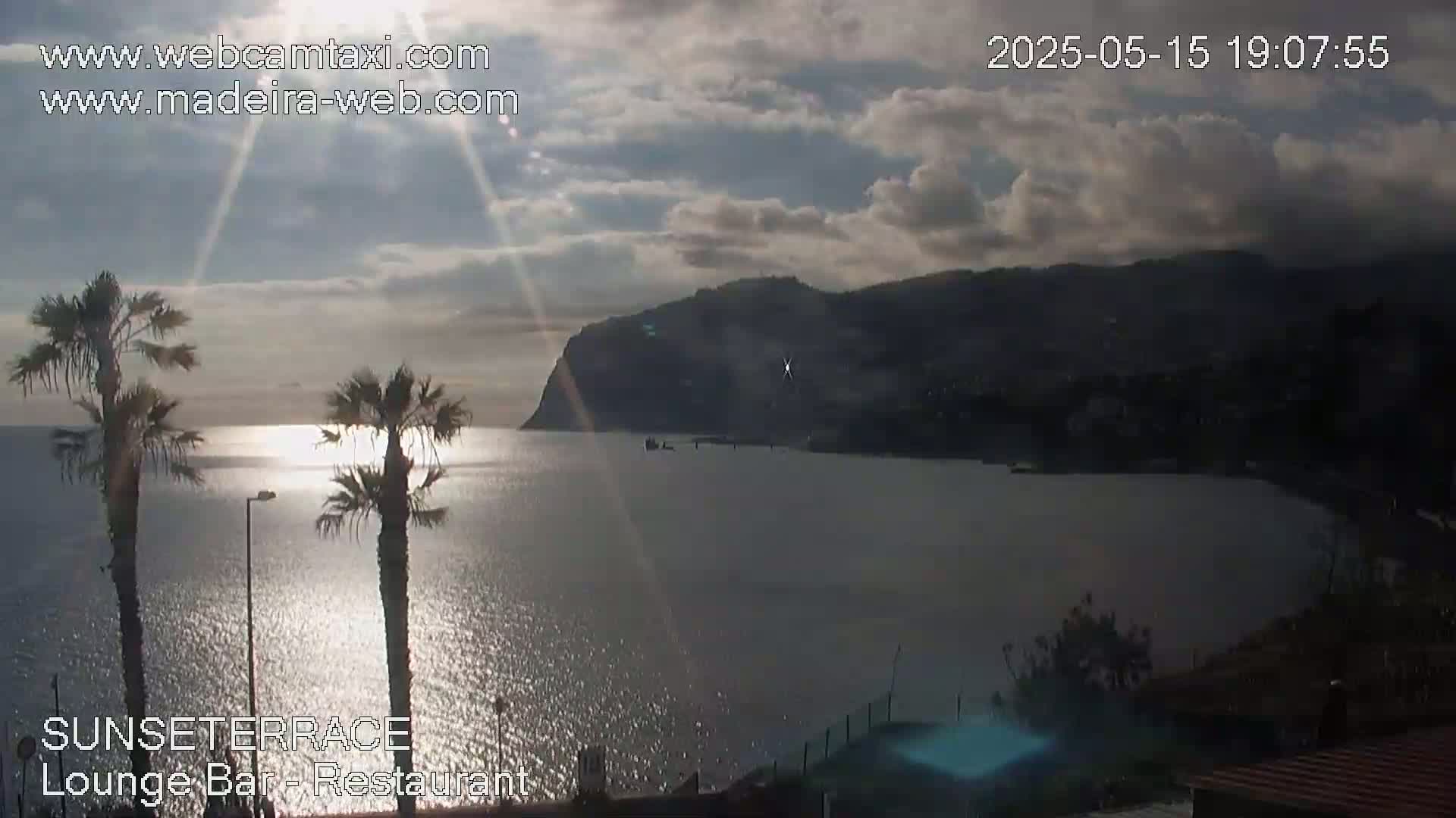 A partly cloudy sunset over a calm body of water, with palm trees in the foreground and a large, dark mountain in the distance.