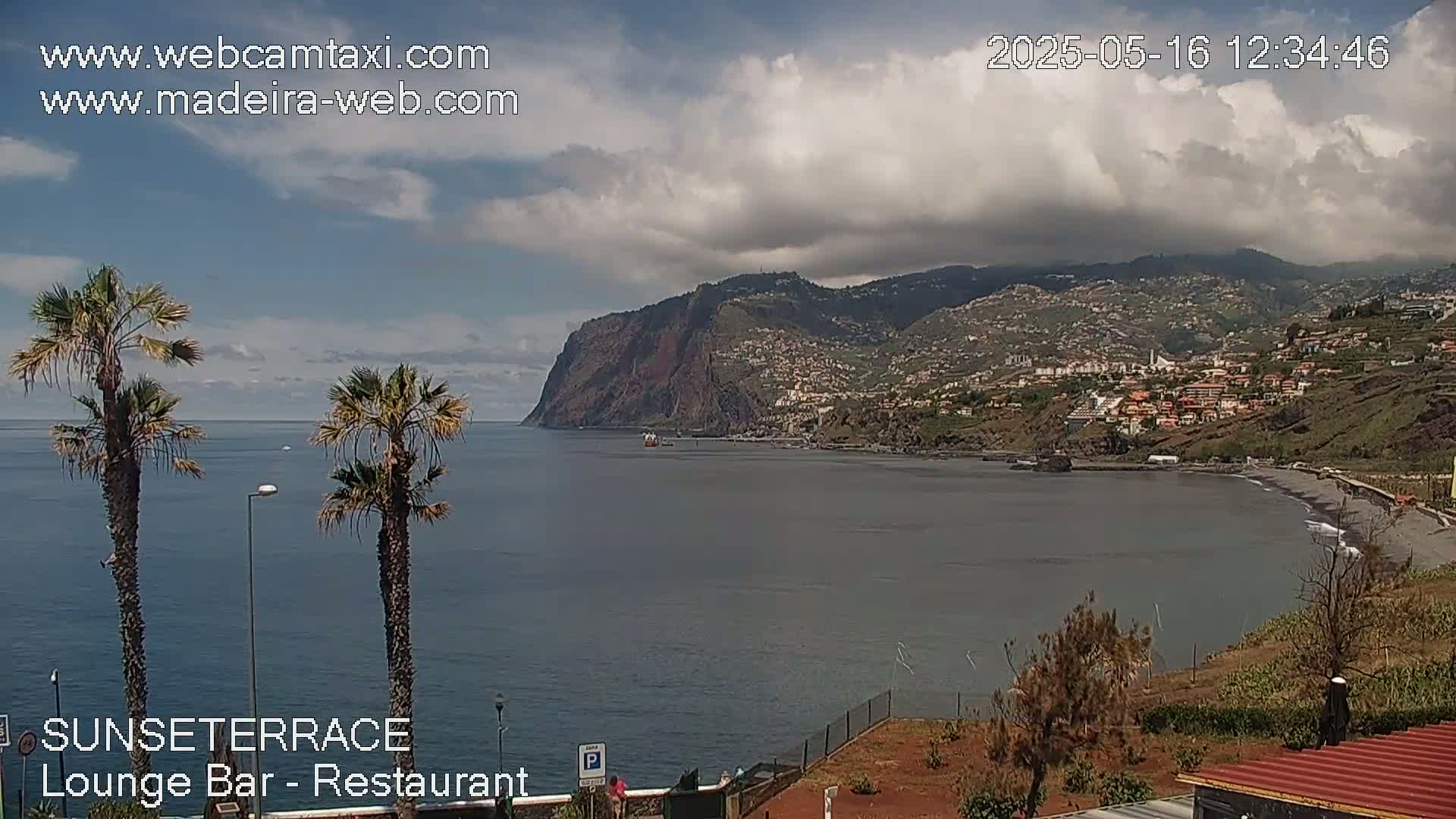 A partly cloudy day reveals a coastal scene with a dark-colored beach, a calm ocean, and a large, mountainous cliffside town in the distance, framed by two palm trees in the foreground.