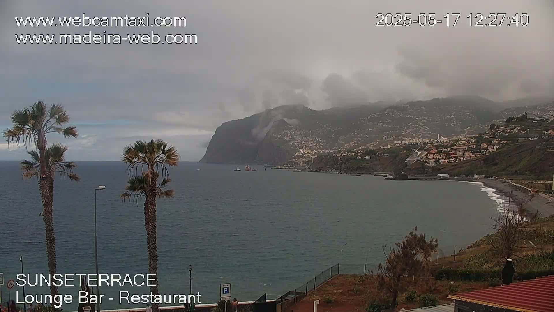 A coastal scene on an overcast day shows a dark-grey ocean, a dark-colored beach, and a hillside town under a cloudy sky.