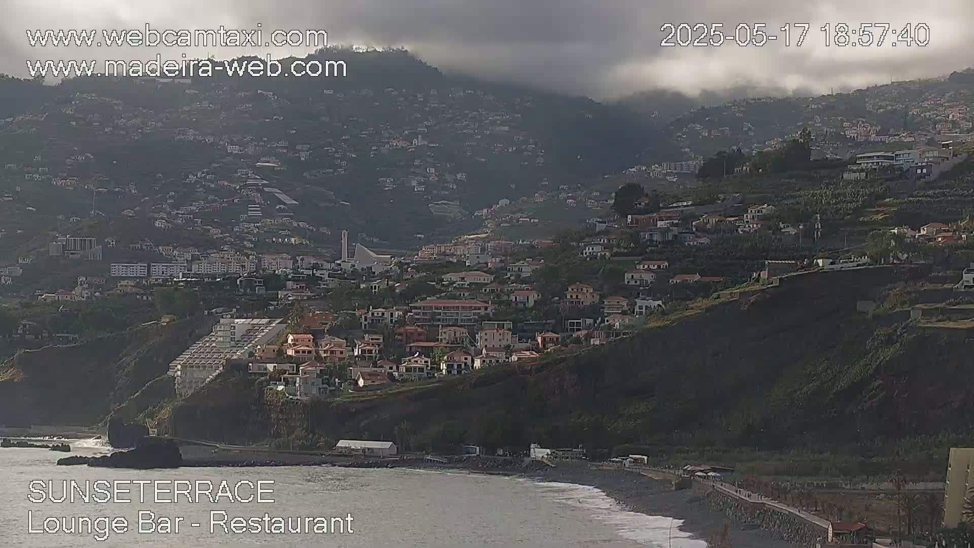 A coastal town on a hillside is partially obscured by clouds under an overcast sky, with a dark ocean visible below.