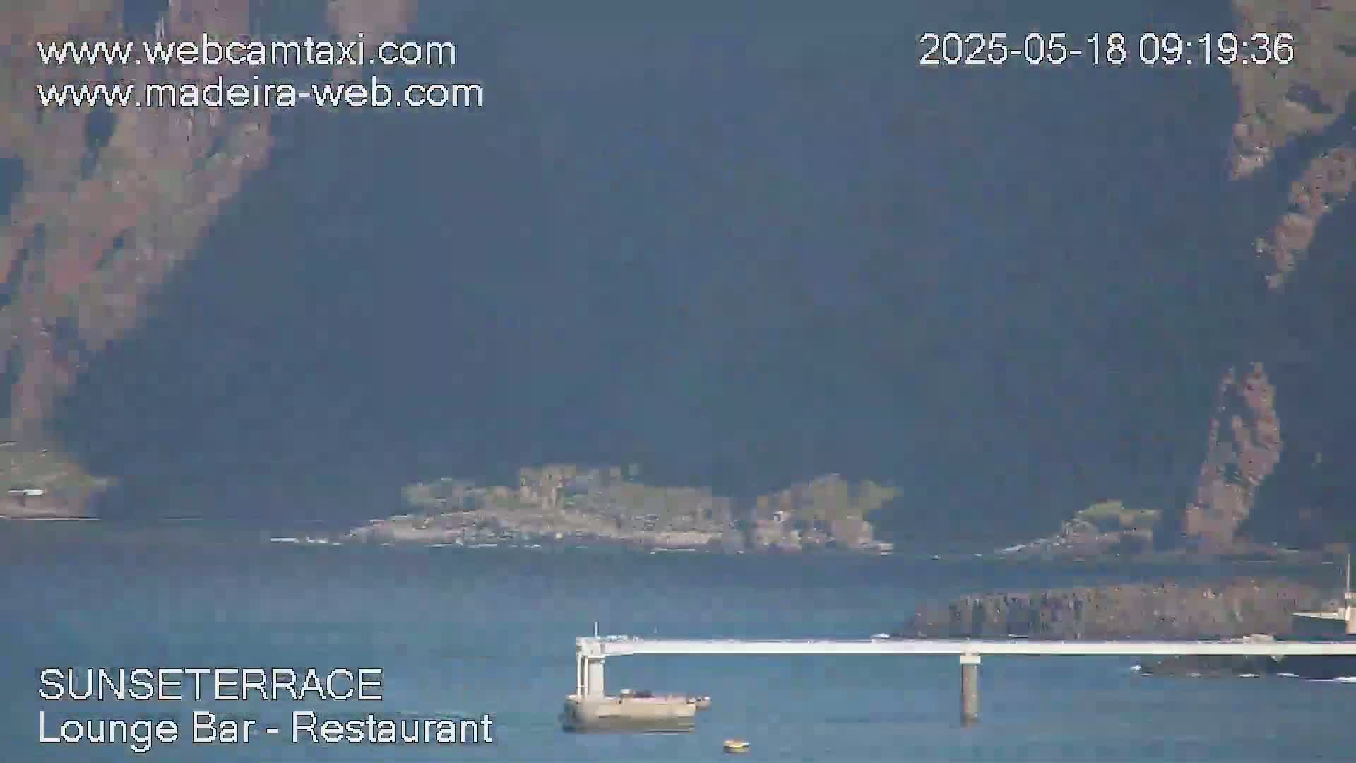 A long, light-colored pier extends over calm blue water, with dark cliffs and land visible in the background under a hazy sky.