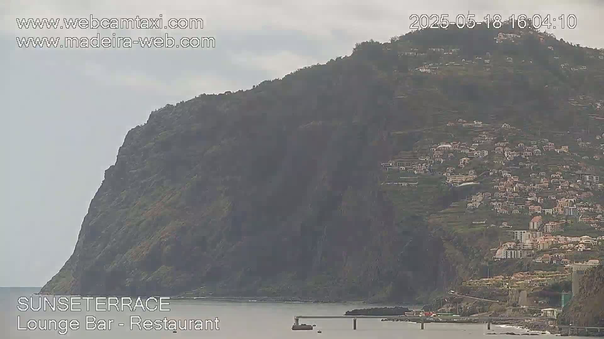 A large, dark-colored cliffside covered in houses and vegetation overlooks a calm body of water under an overcast sky.