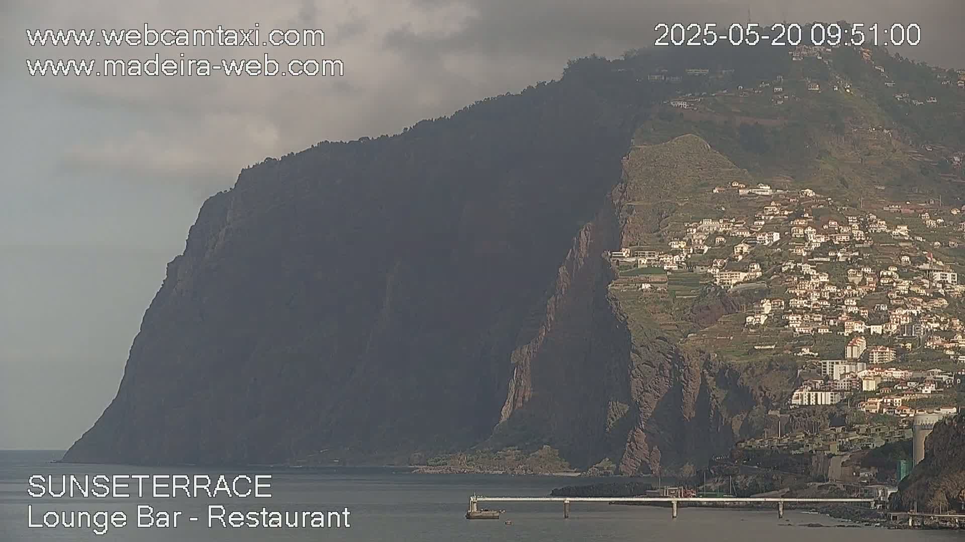 A large, dark cliff overlooks a town built into a hillside, all under an overcast sky.