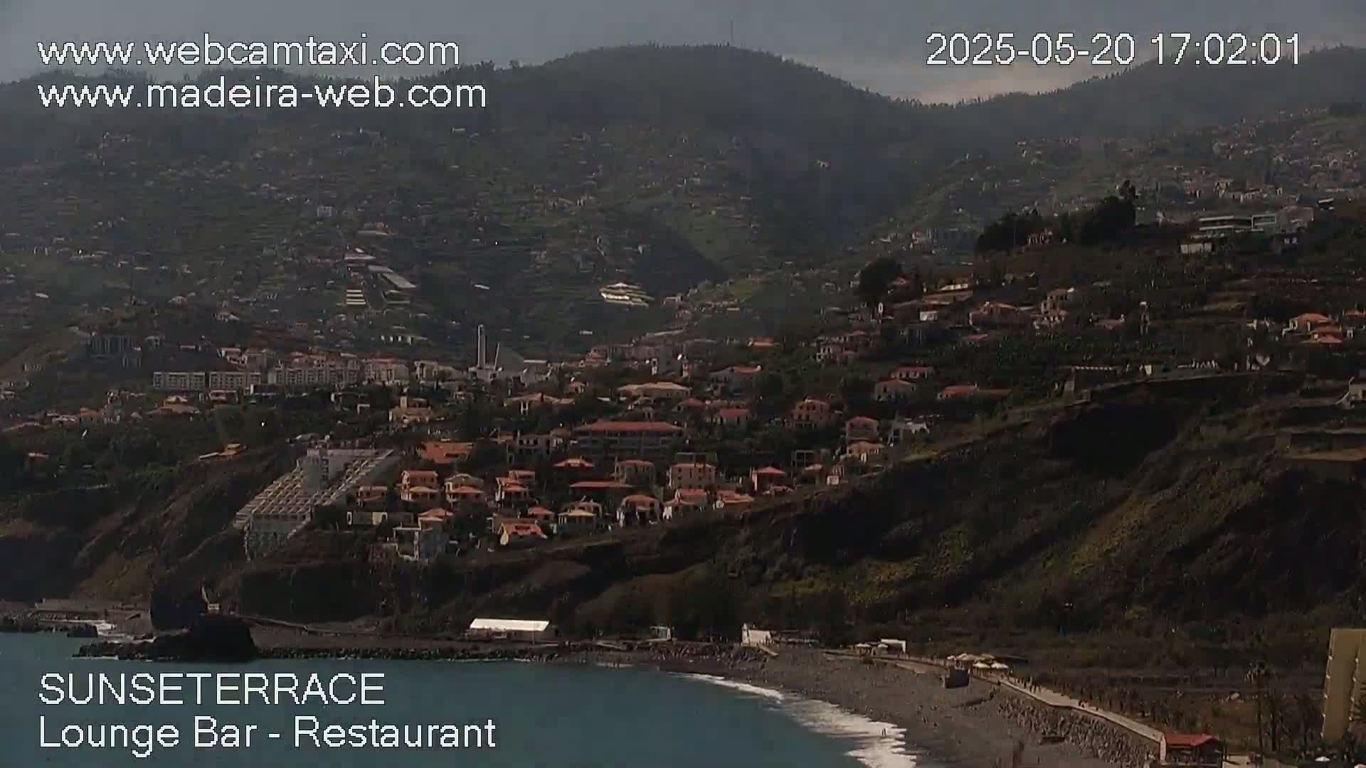 A coastal town nestled on a hillside overlooking a dark-sand beach and ocean under hazy, daytime conditions.