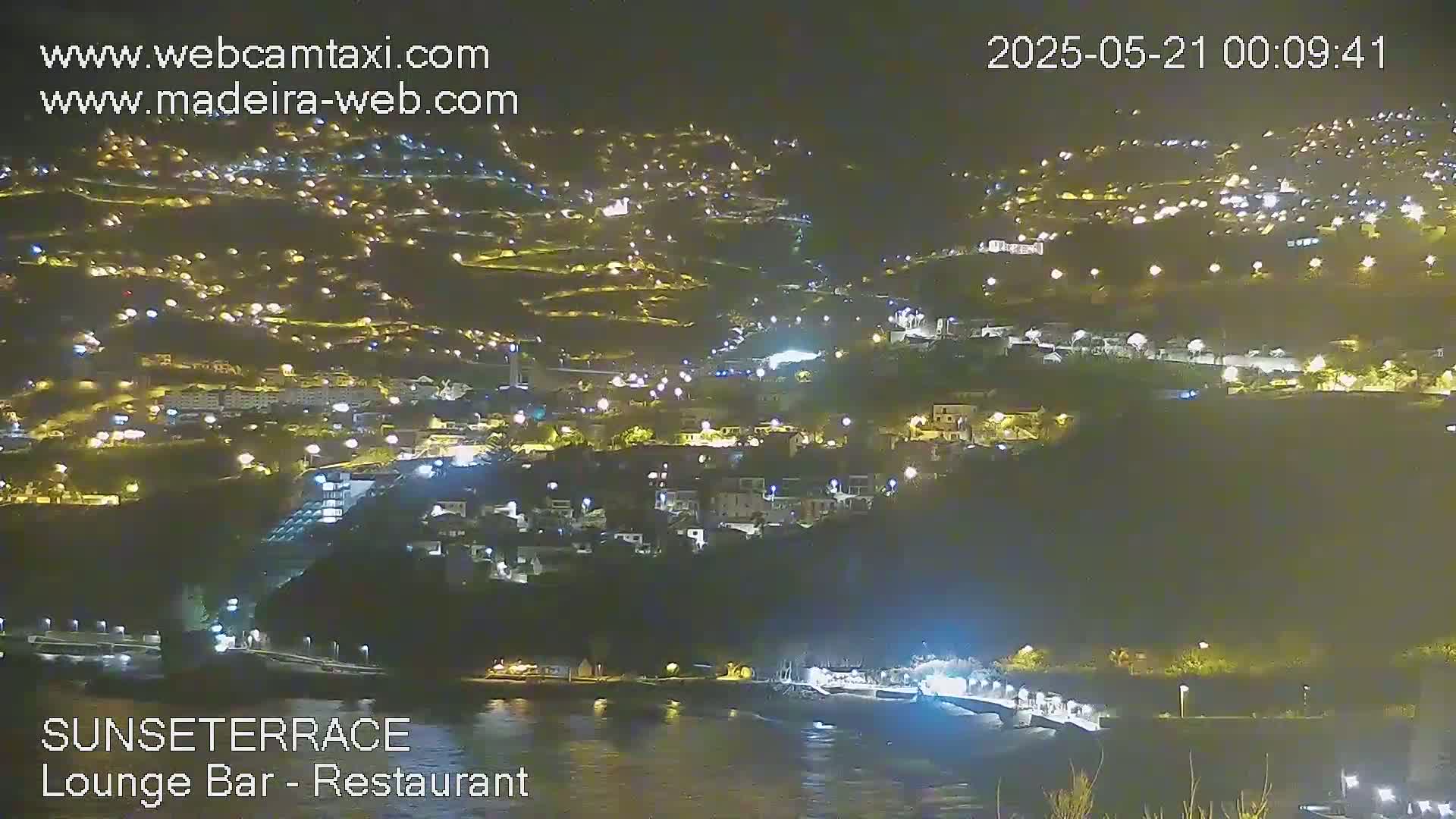 A nighttime, aerial view shows a hillside town illuminated by many lights, next to dark water.