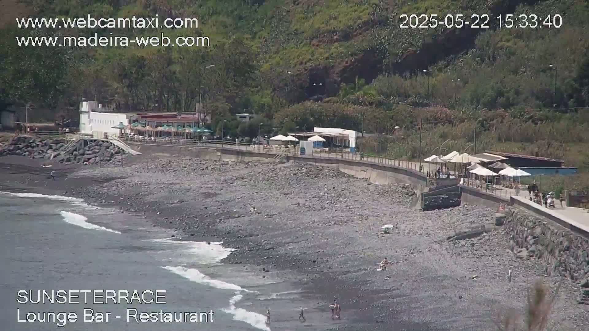 A rocky beach with a restaurant and lounge bar,  a few people, and gentle waves is seen on a sunny day.