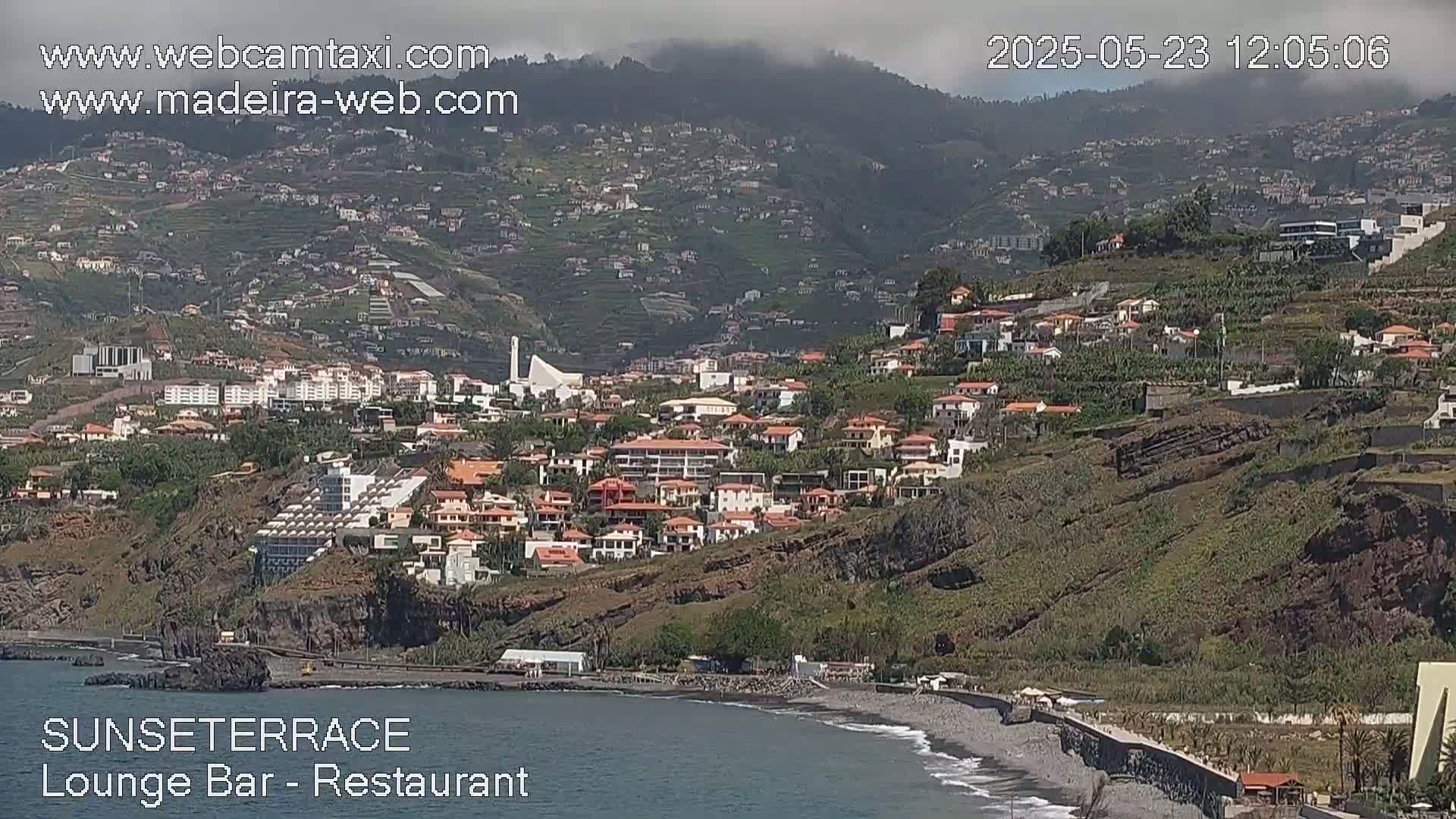 A coastal town nestled on a hillside overlooking a dark-sand beach on a partly cloudy day.