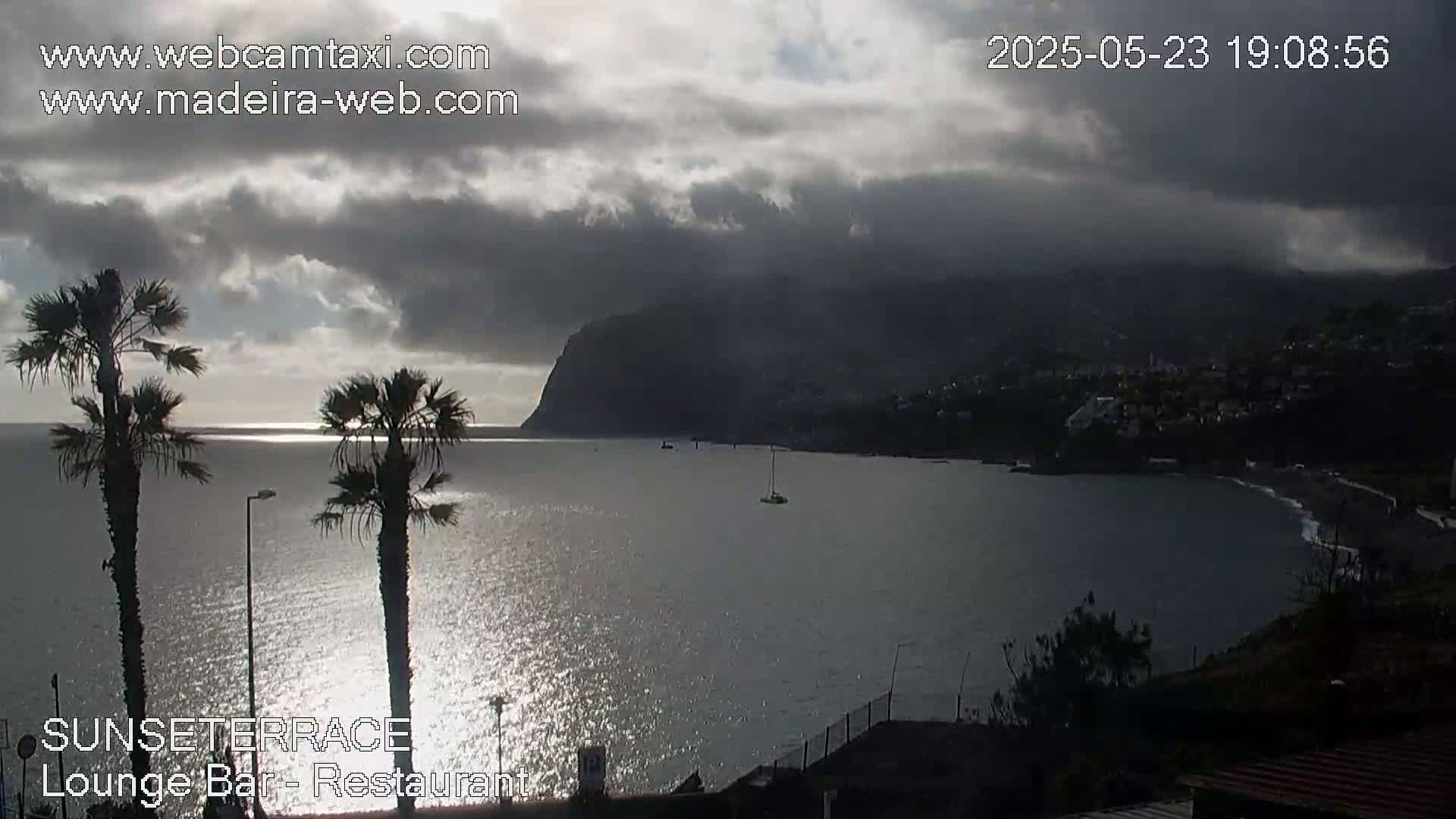 A partly cloudy, late afternoon or early evening scene overlooking a calm bay with a dark mountain in the background, two palm trees in the foreground, and a few boats on the water.