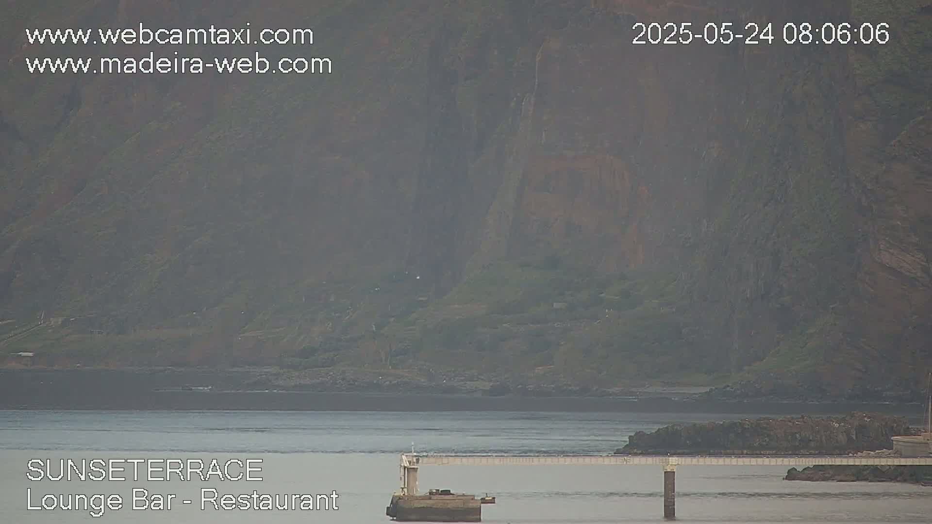 A hazy view shows a rocky cliffside overlooking a calm body of water with a long pier extending from the shore.