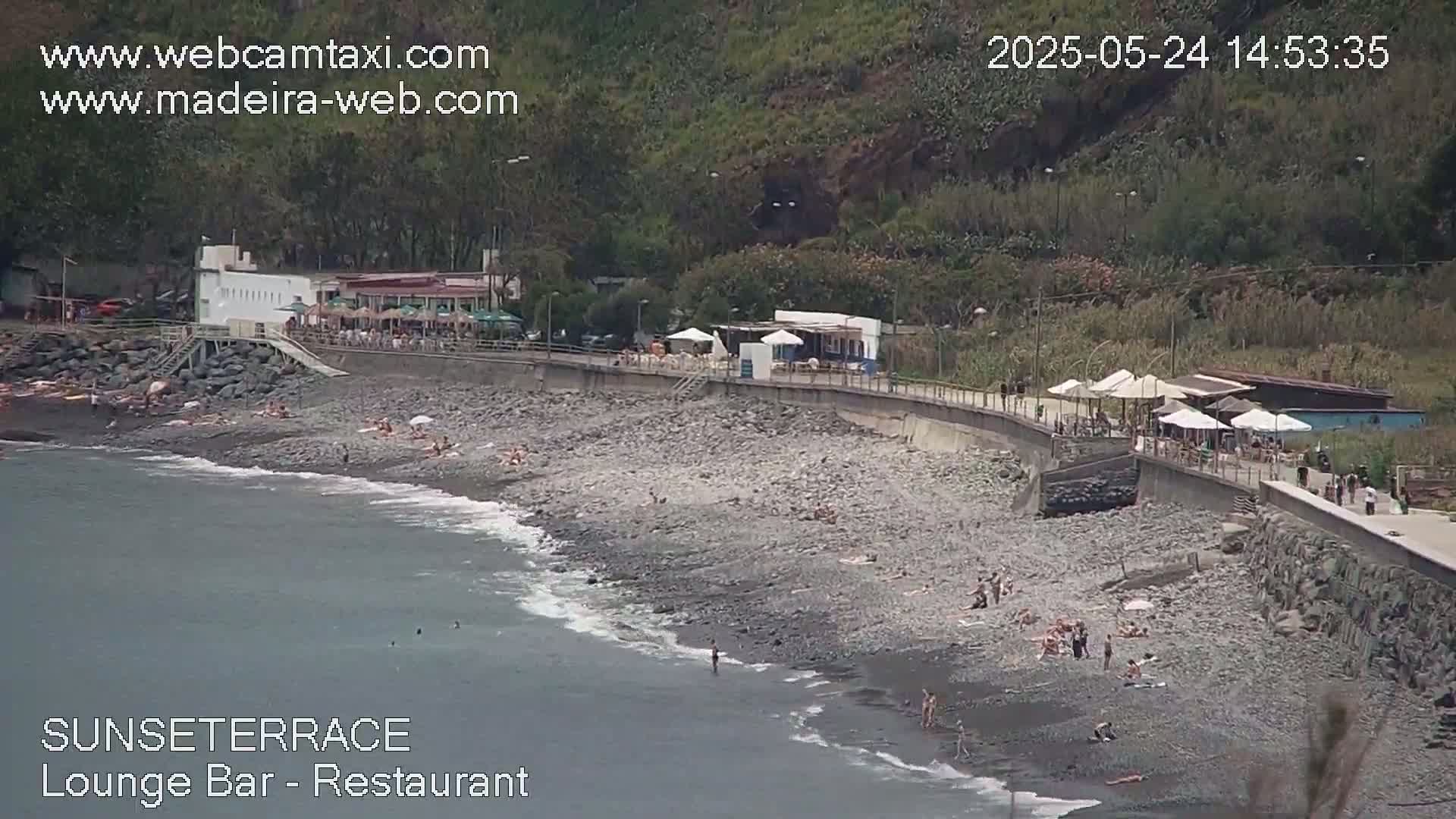 A rocky beach with a restaurant and several people sunbathing or swimming, situated below a hillside on a partly cloudy day.