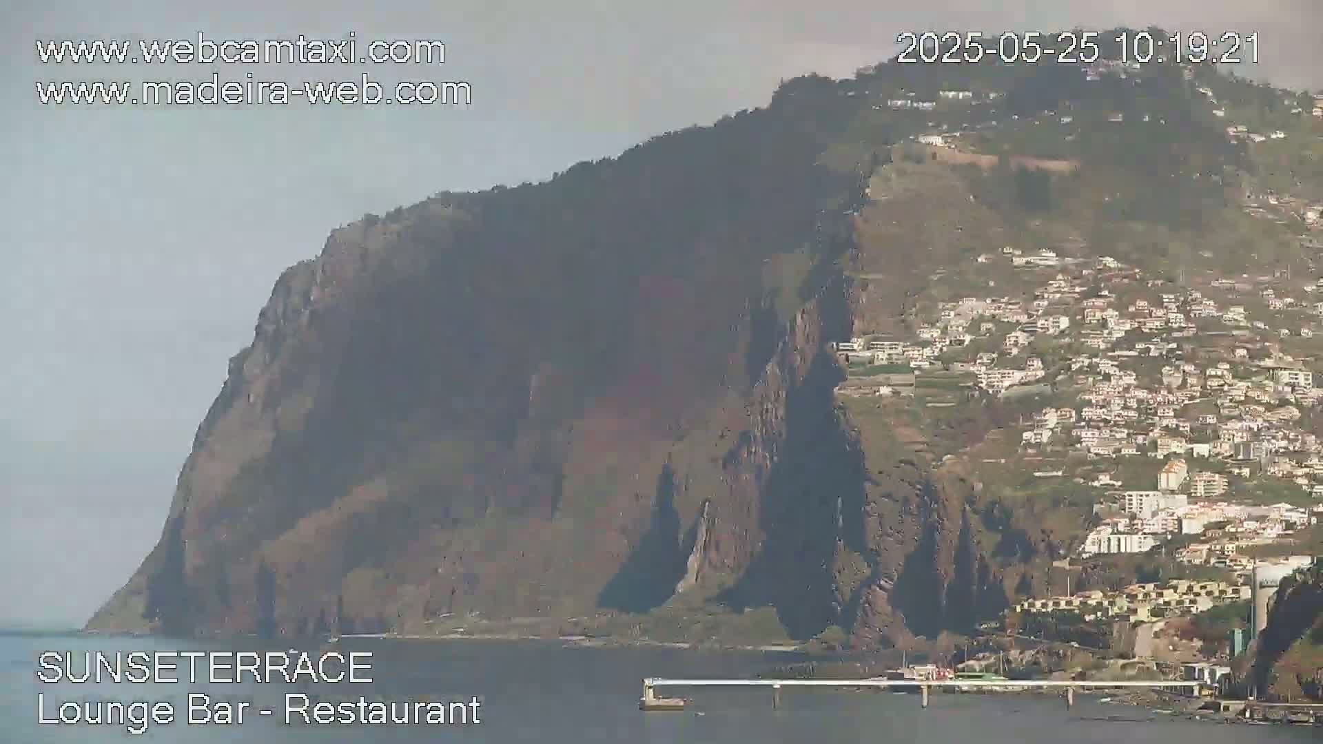 A hazy view shows a large, dark cliff overlooking a town nestled on a hillside that slopes down to a calm body of water.
