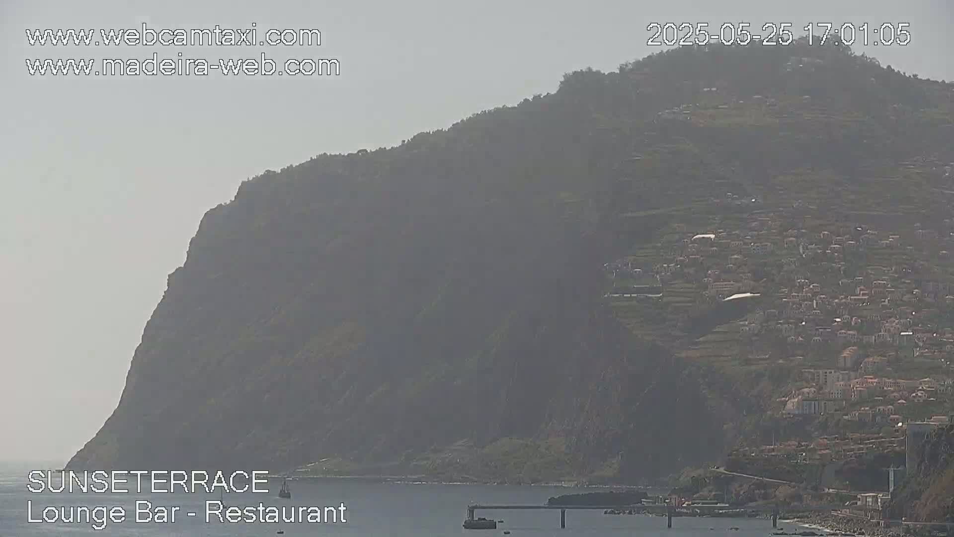 A hazy view shows a coastal town nestled on a hillside overlooking a calm body of water, with a large cliff face dominating the left side of the scene.