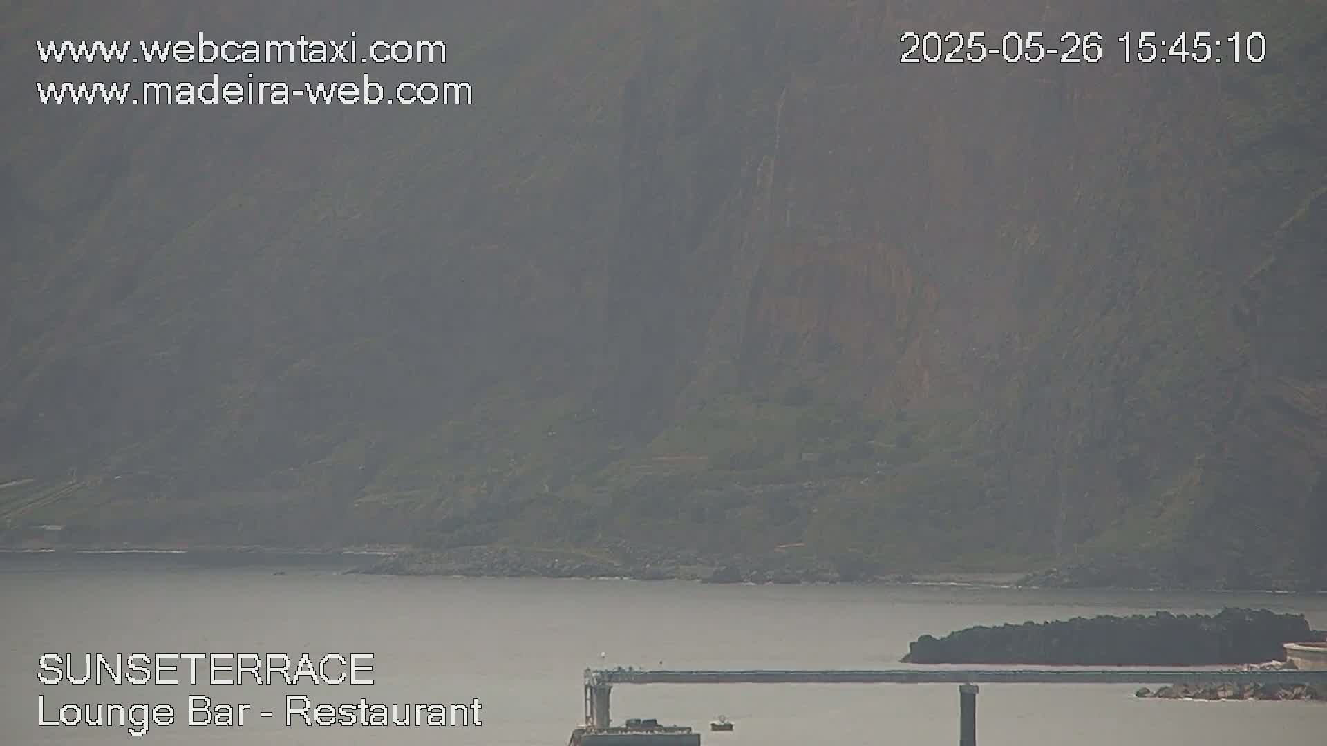 A hazy view shows a body of water with a long, dark structure extending across it near a rocky shoreline, backed by a dark green hillside.