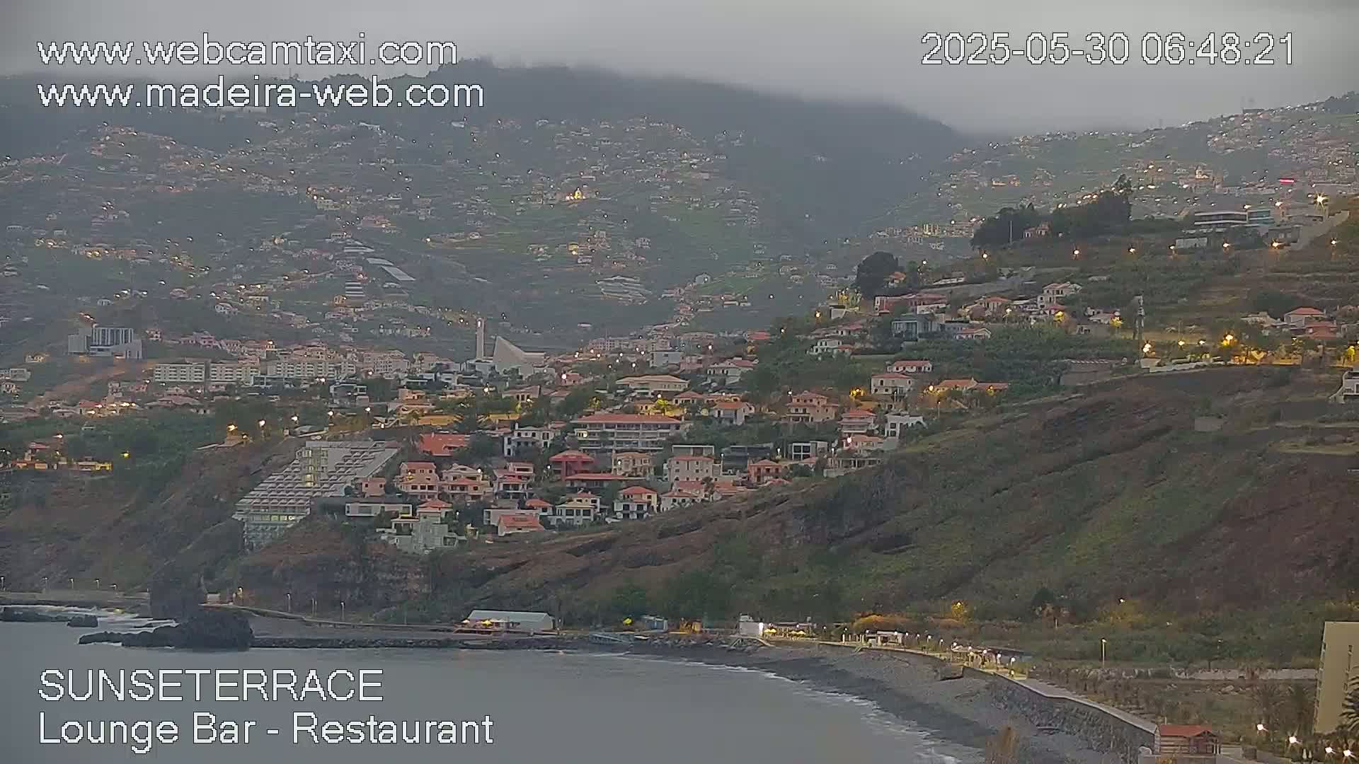 A coastal town built on a hillside, dimly lit at twilight under an overcast sky, stretches down to a dark-sand beach next to a calm ocean.