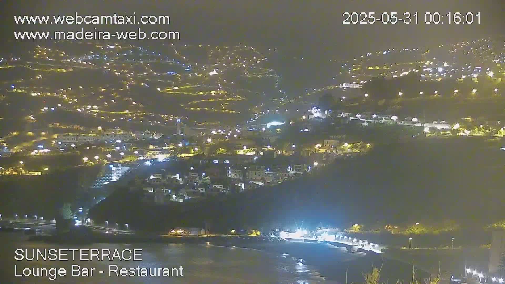 A nighttime, long shot of a hillside town illuminated by numerous lights, overlooking a dark body of water.