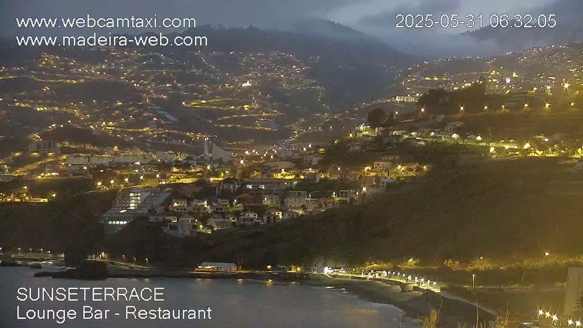 A nighttime coastal town on a hillside is illuminated by numerous lights, under a dark, overcast sky.