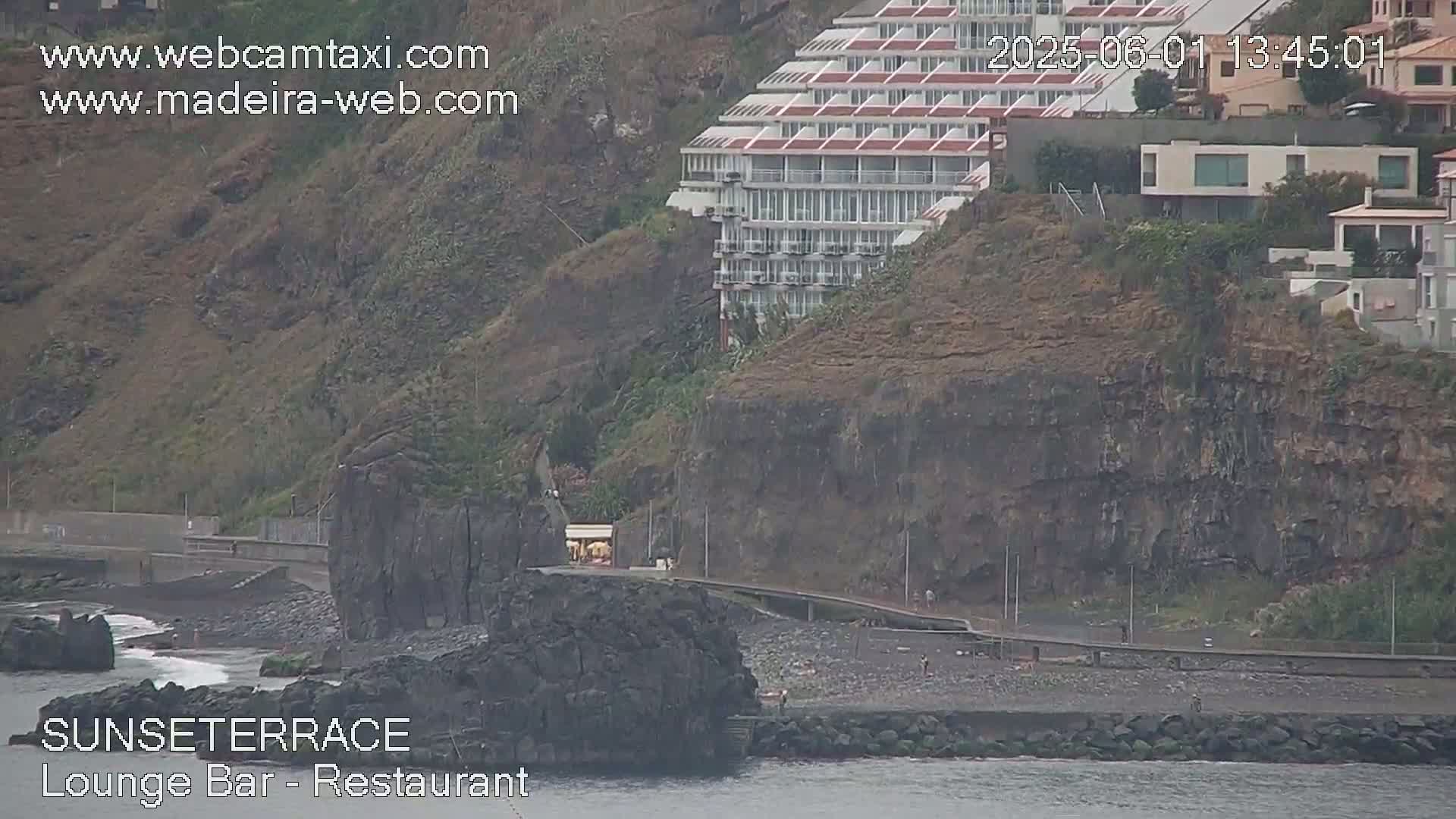 A dark, rocky coastline with a multi-story building on a cliff overlooking a dark-sand beach on an overcast day.