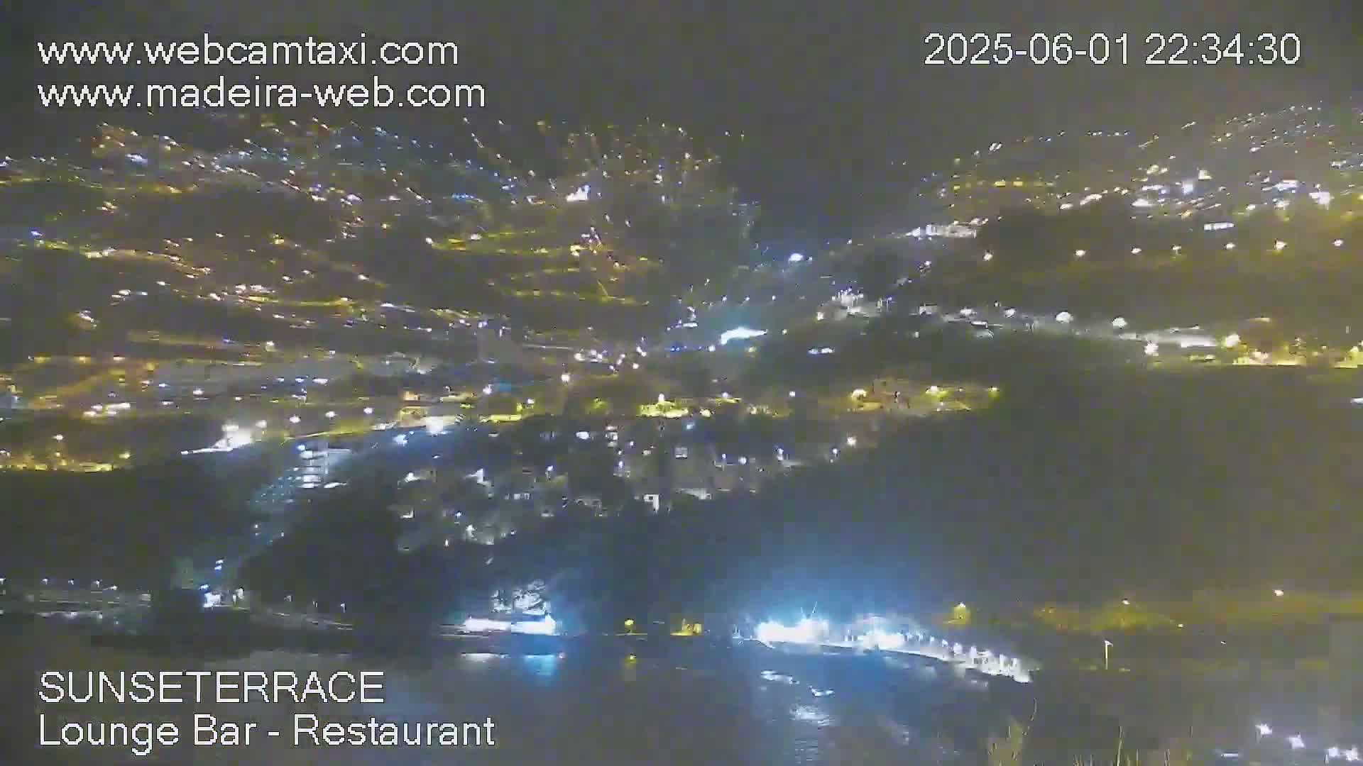 A nighttime, high-angle view of a town illuminated by many lights, situated on a hillside near a body of water.