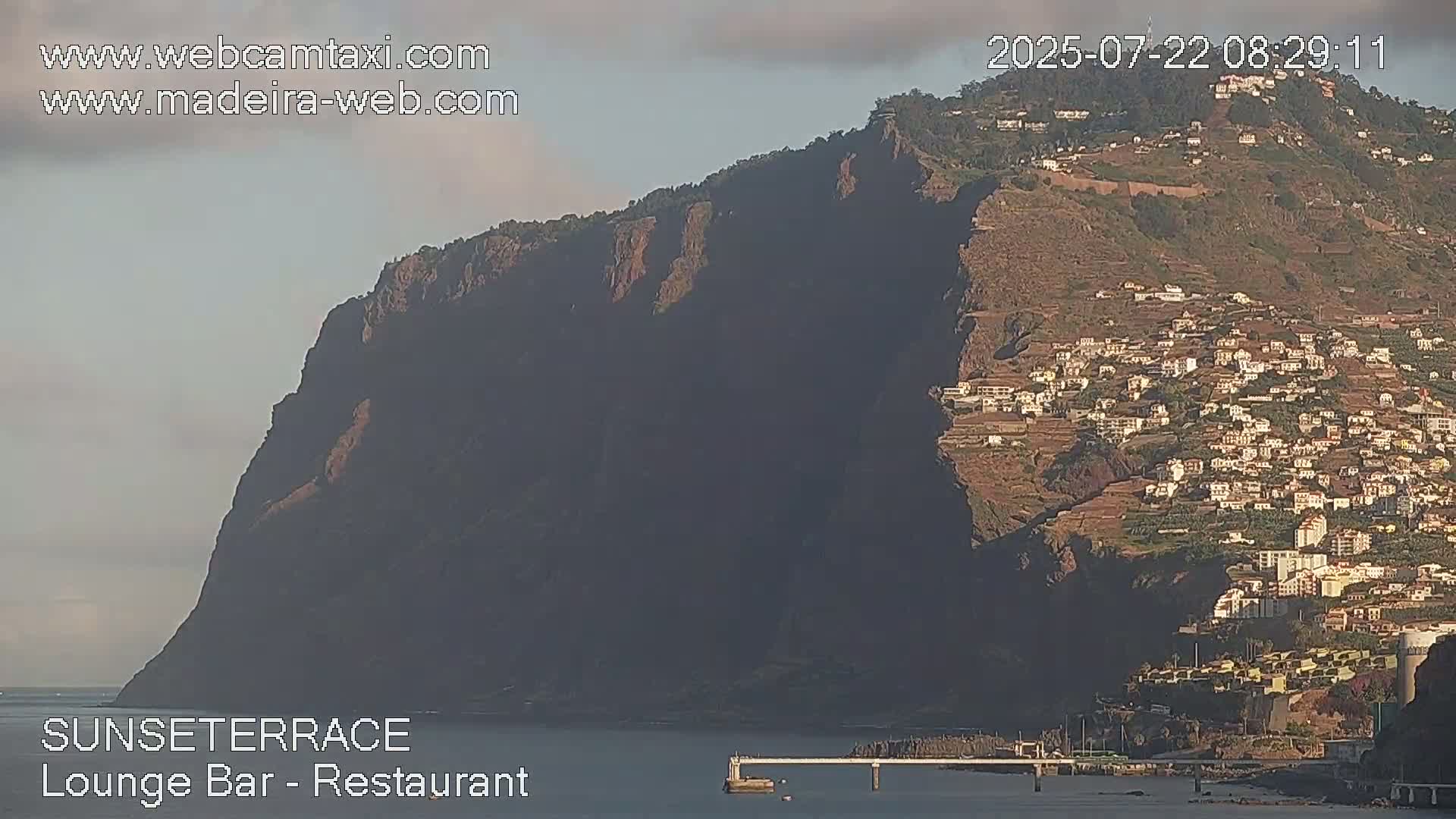 A large, dark cliff overlooks a town nestled on its slopes, with calm water visible at the base under a partly cloudy sky.