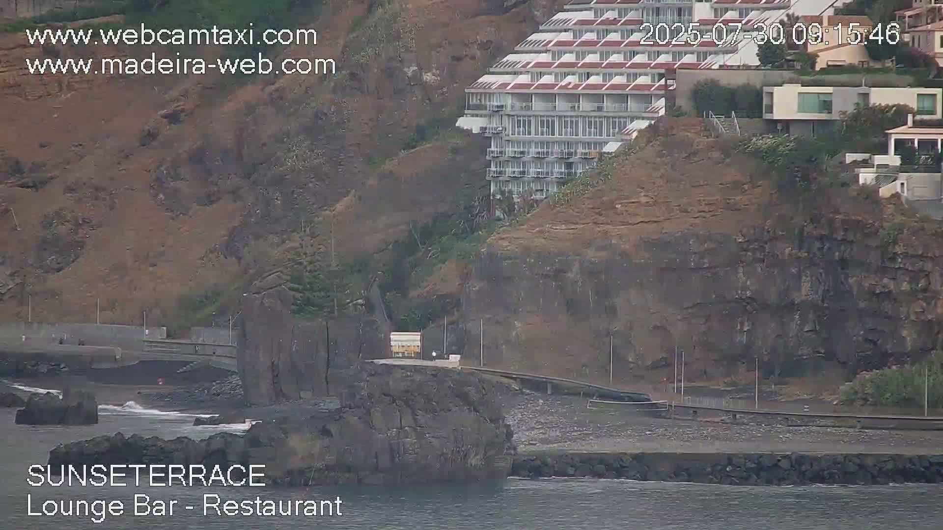A seaside cliffside features a multi-story hotel, a dark-colored beach, and a rocky outcropping, all under an overcast sky.