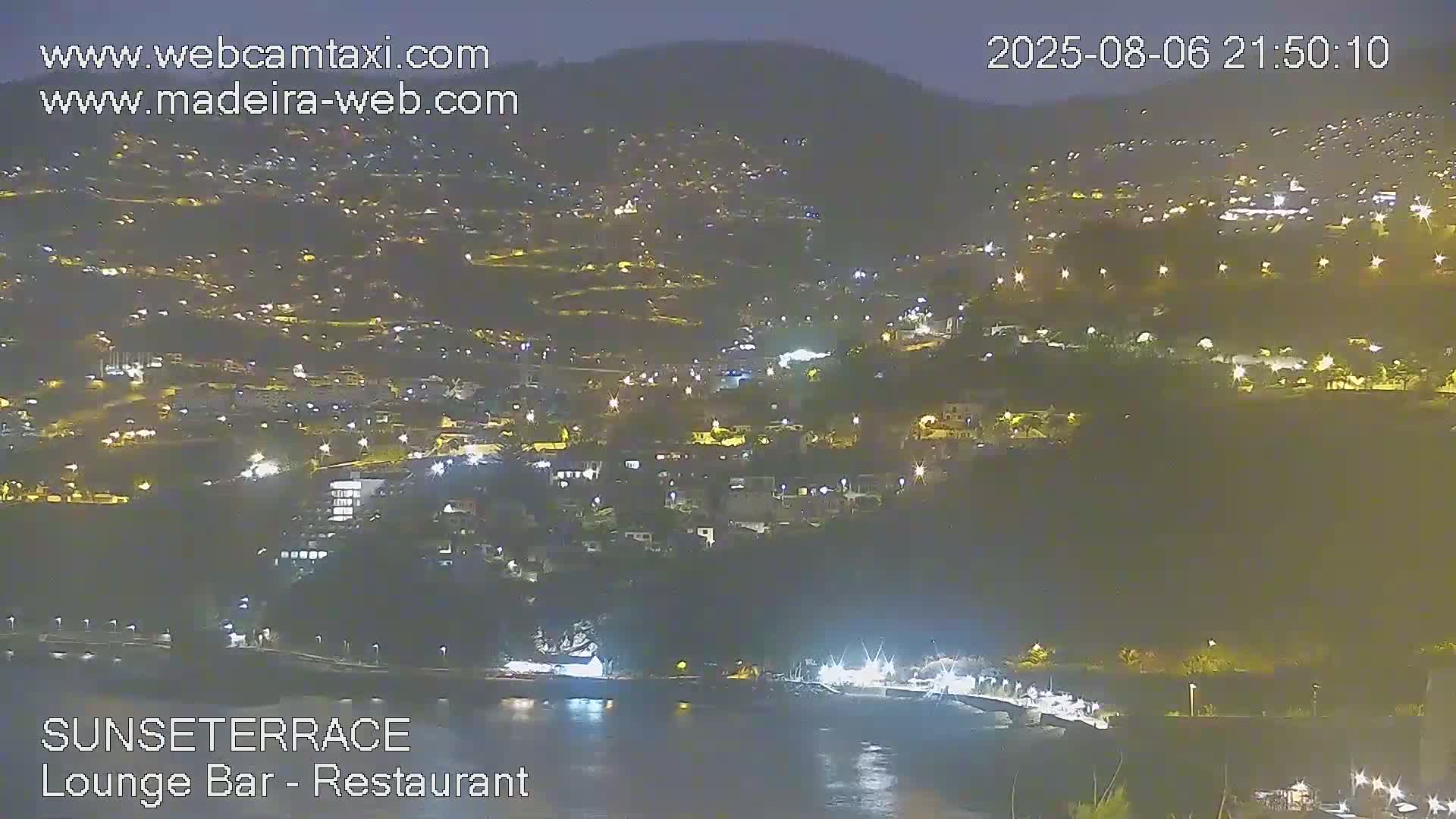 A nighttime view shows a hillside town illuminated by lights, extending down to a dark body of water with a brightly lit waterfront.