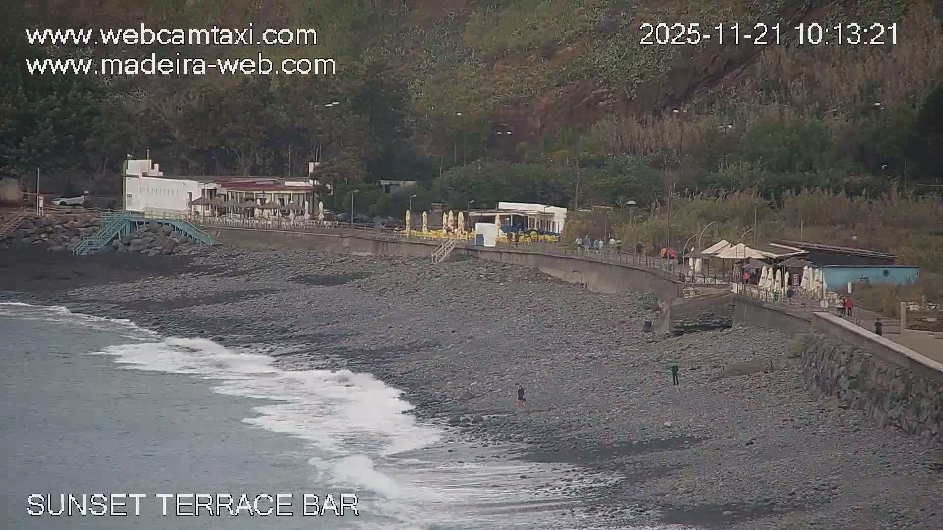 On a clear and sunny day, ocean waves lap a wide, rocky beach, bordered by a promenade lined with restaurants and bars offering outdoor seating, with a few people scattered along the shore and path, all framed by a lush, green hillside.