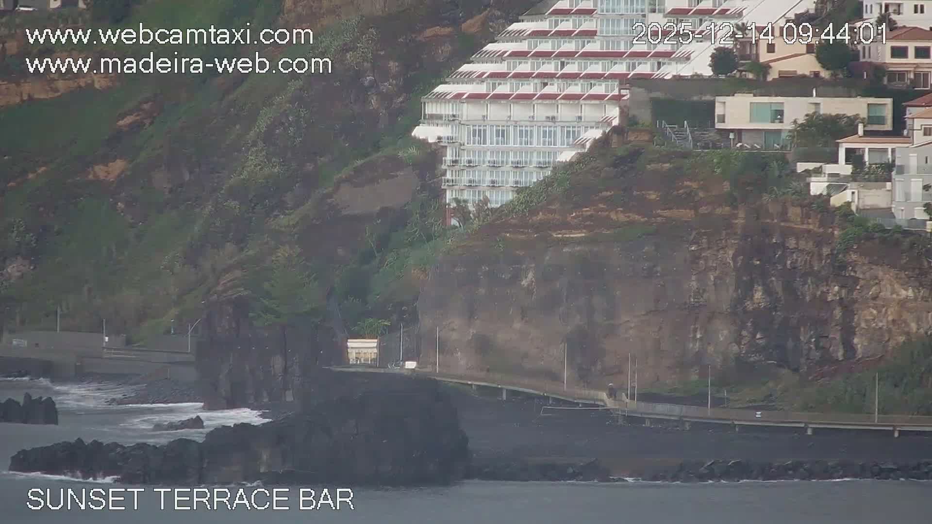 On a clear and sunny day, ocean waves lap a wide, rocky beach, bordered by a promenade lined with restaurants and bars offering outdoor seating, with a few people scattered along the shore and path, all framed by a lush, green hillside.