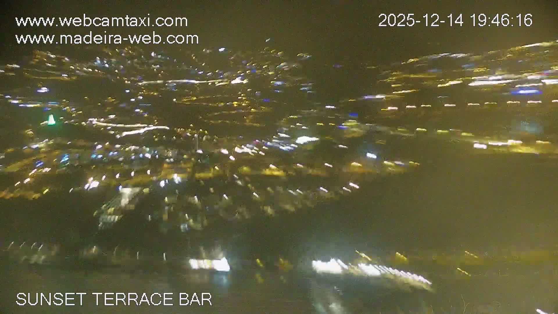 On a clear and sunny day, ocean waves lap a wide, rocky beach, bordered by a promenade lined with restaurants and bars offering outdoor seating, with a few people scattered along the shore and path, all framed by a lush, green hillside.