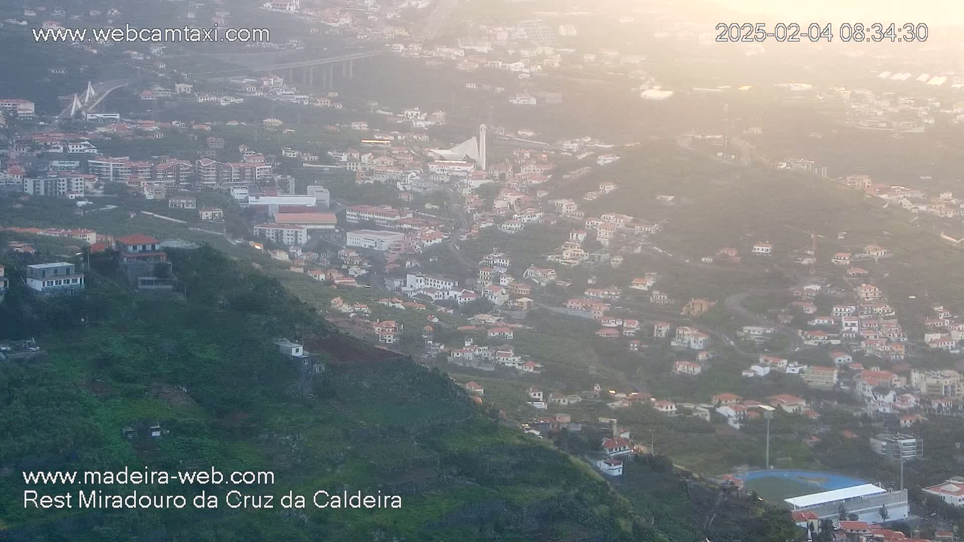 A hazy, high-angle view shows a hillside town with many white buildings, a large sports field, and a bridge in the distance.