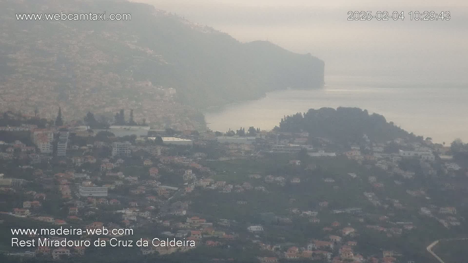 A hazy view shows a coastal town nestled between hills and a calm body of water on a foggy day.