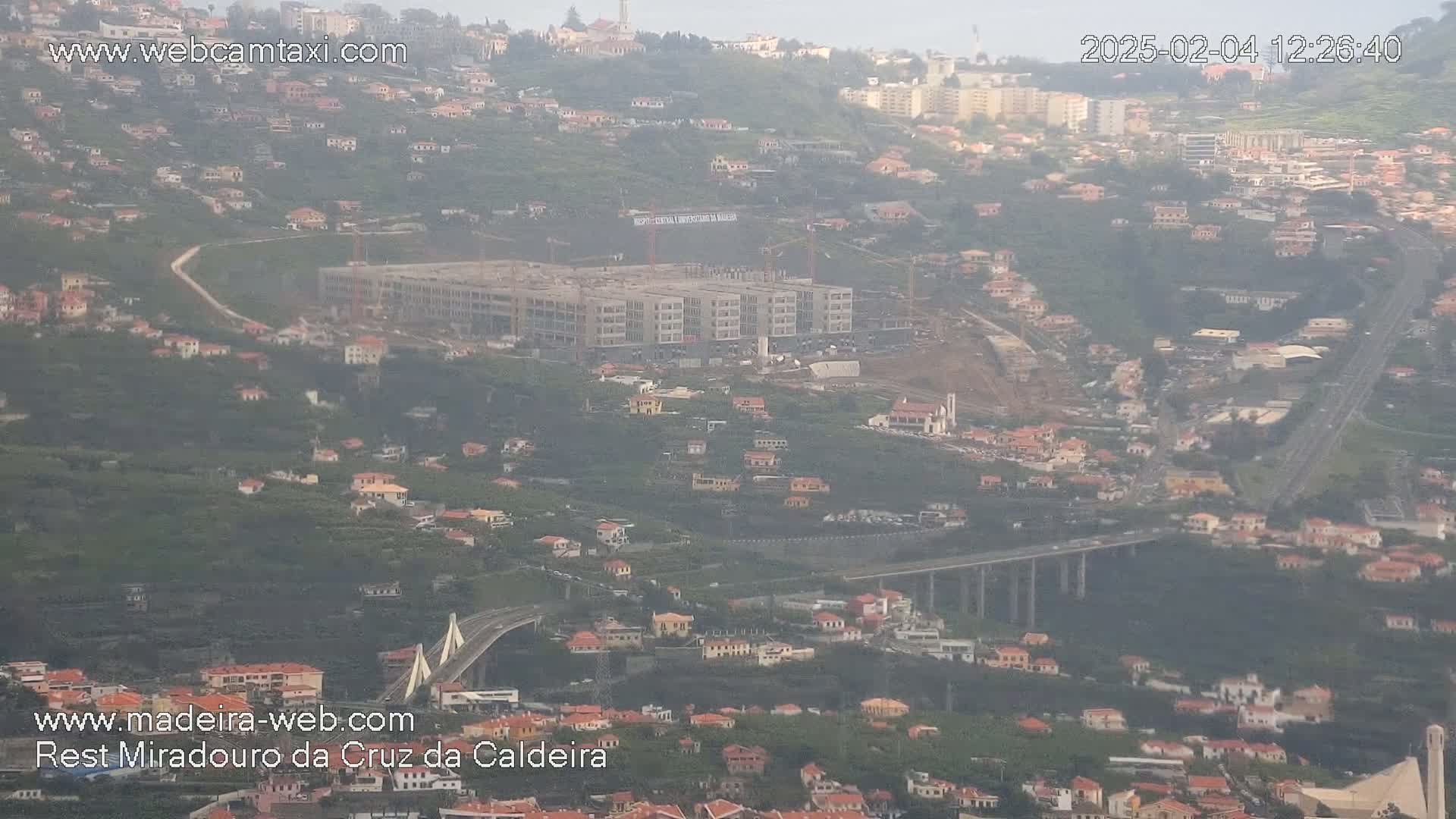 A hazy view of a hillside town with a partially-constructed multi-story building and a bridge, under a cloudy sky.