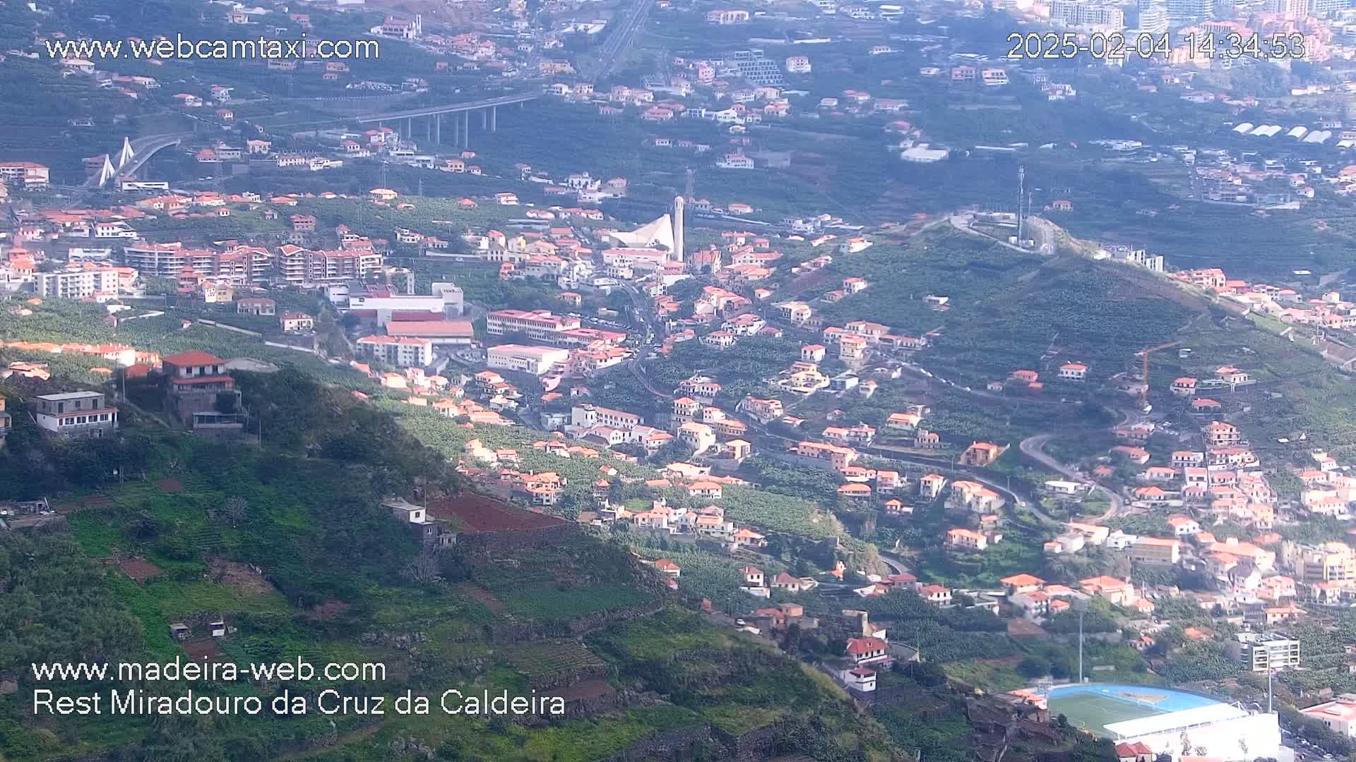 An aerial view shows a hillside town with numerous low, orange-roofed buildings nestled amongst terraced greenery, a bridge in the distance, and a hazy, bright sky.