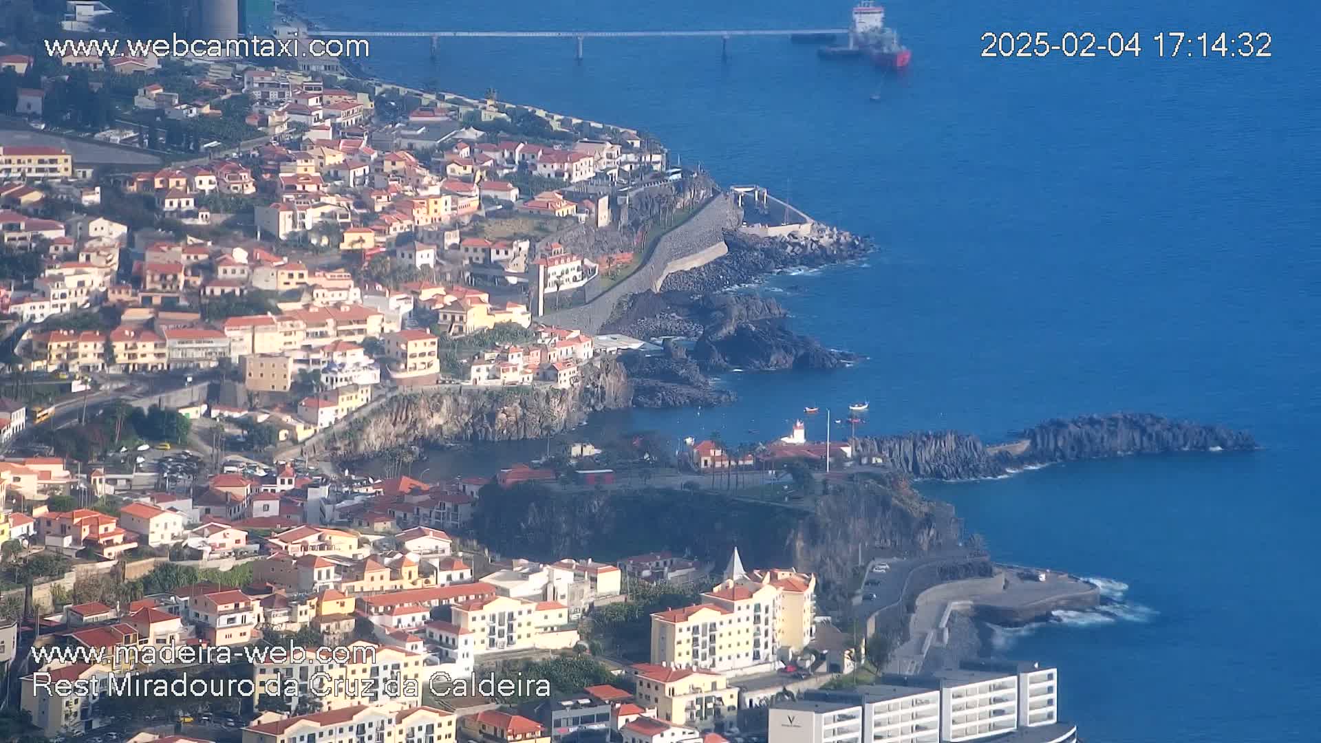 An aerial view shows a coastal town with many low buildings, a rocky coastline, and a calm blue ocean under a sunny sky.