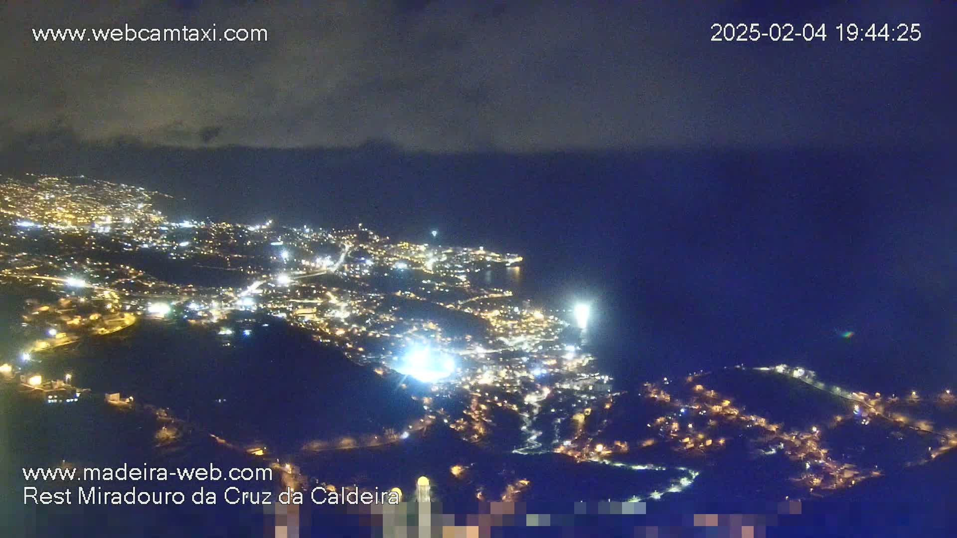 A nighttime, high-angle view of a coastal town illuminated by lights, extending down a hillside toward a dark body of water under a mostly clear sky.