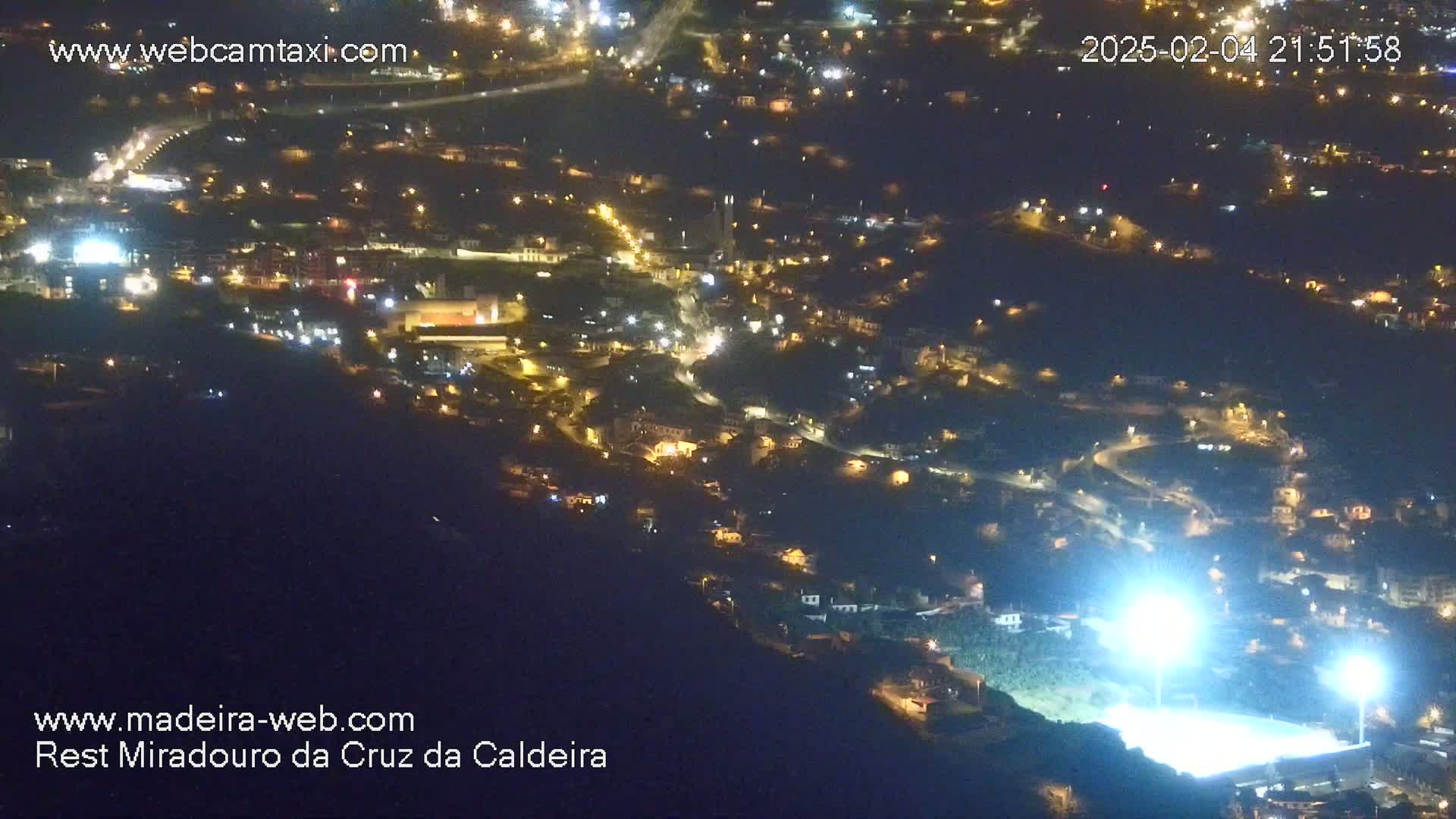 A nighttime, high-angle view shows a brightly lit sports field and numerous houses with illuminated windows spread across a hillside.