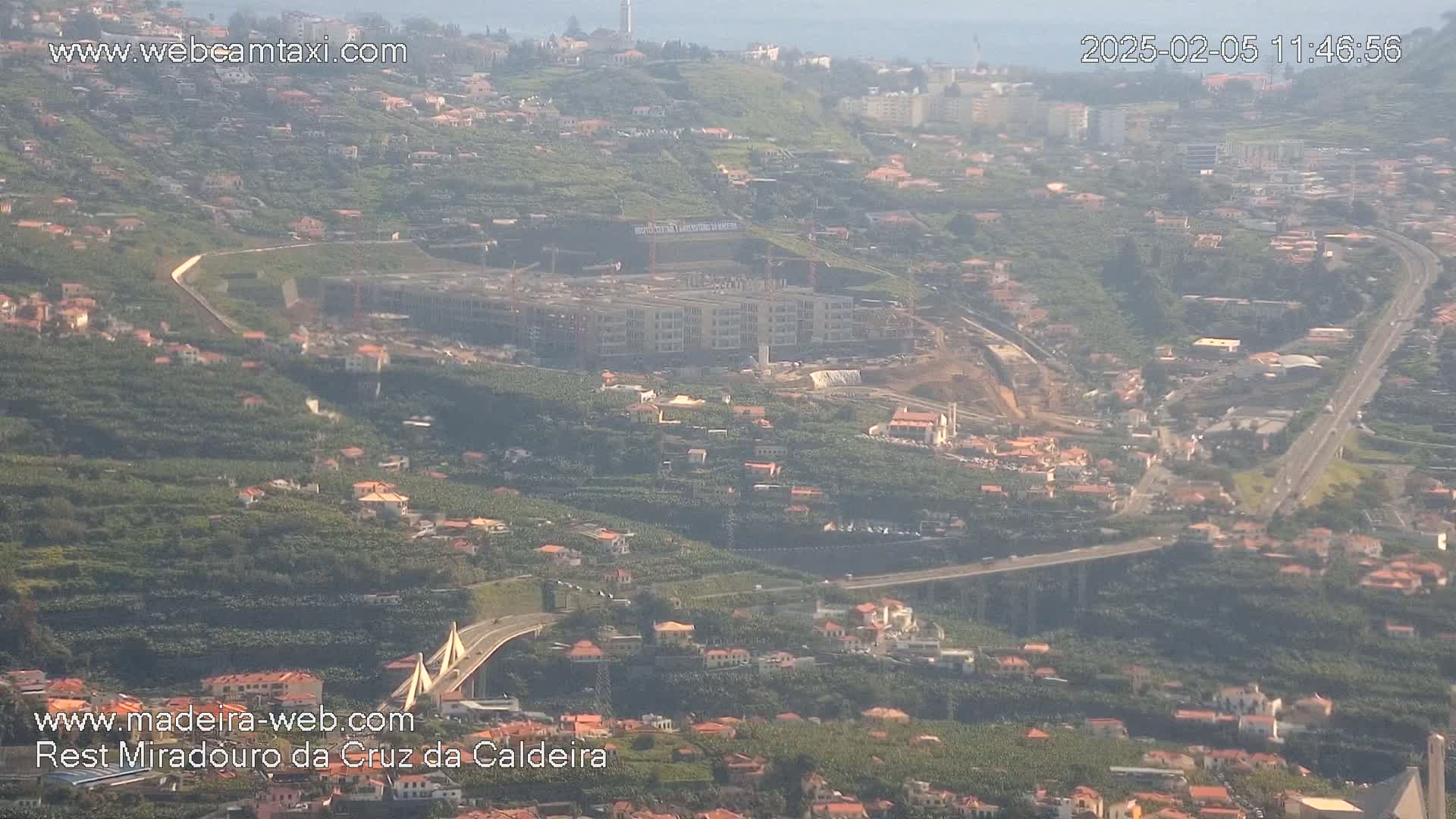 A hazy aerial view shows a coastal town with a large construction site, a bridge, and numerous buildings nestled among lush green hillsides.