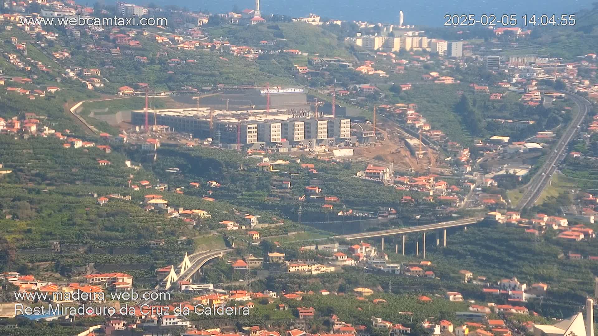 A high-angle view of a hillside town with a large building under construction, a highway bridge, and lush green vegetation on a sunny day.