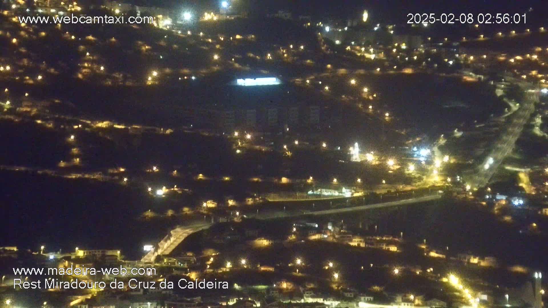 A nighttime, aerial view shows a town illuminated by numerous lights, scattered across a hilly landscape.