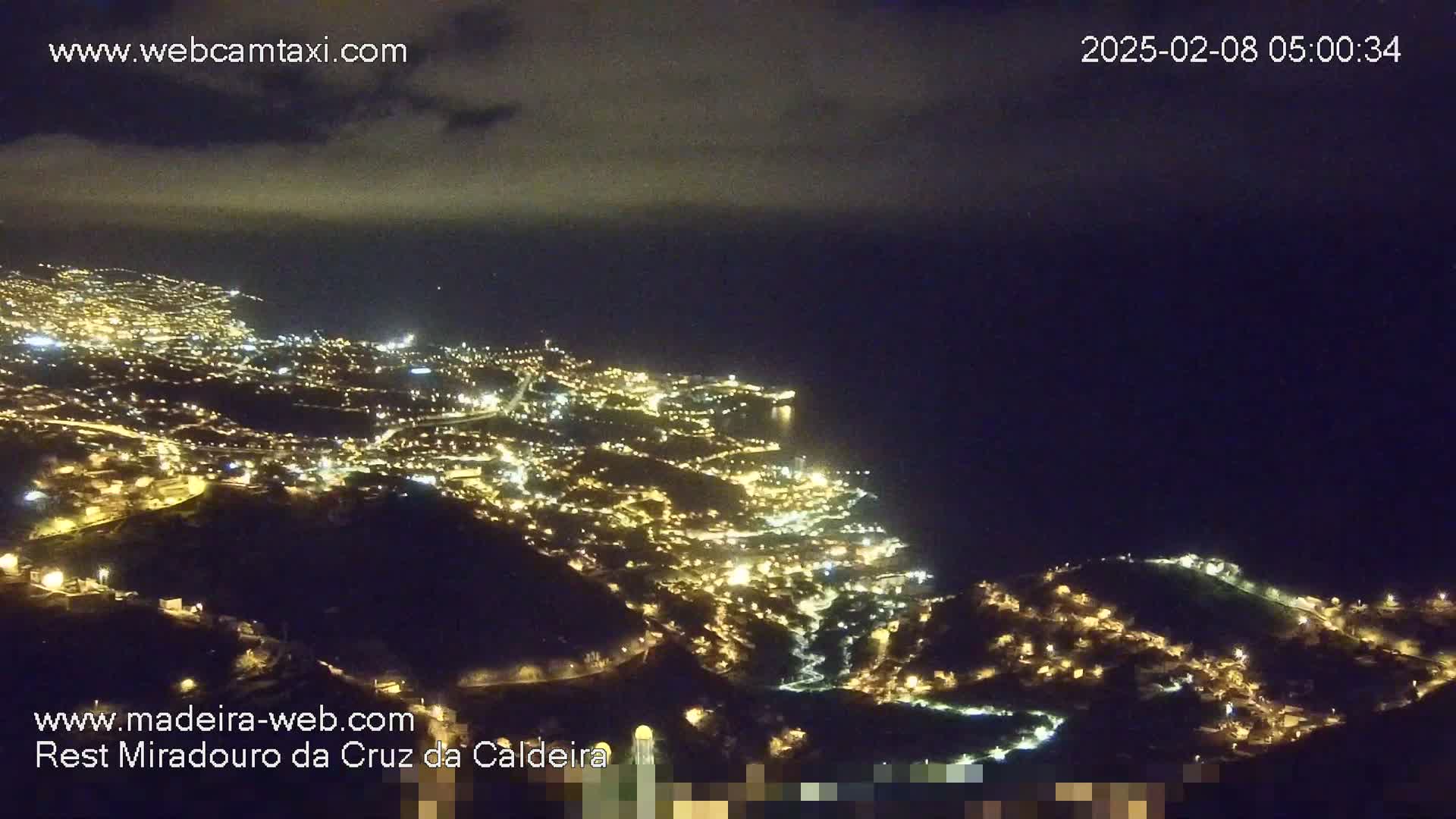 A nighttime, high-angle view shows a coastal town illuminated with many lights, extending along a dark hillside that slopes down to a dark ocean under a mostly clear sky.