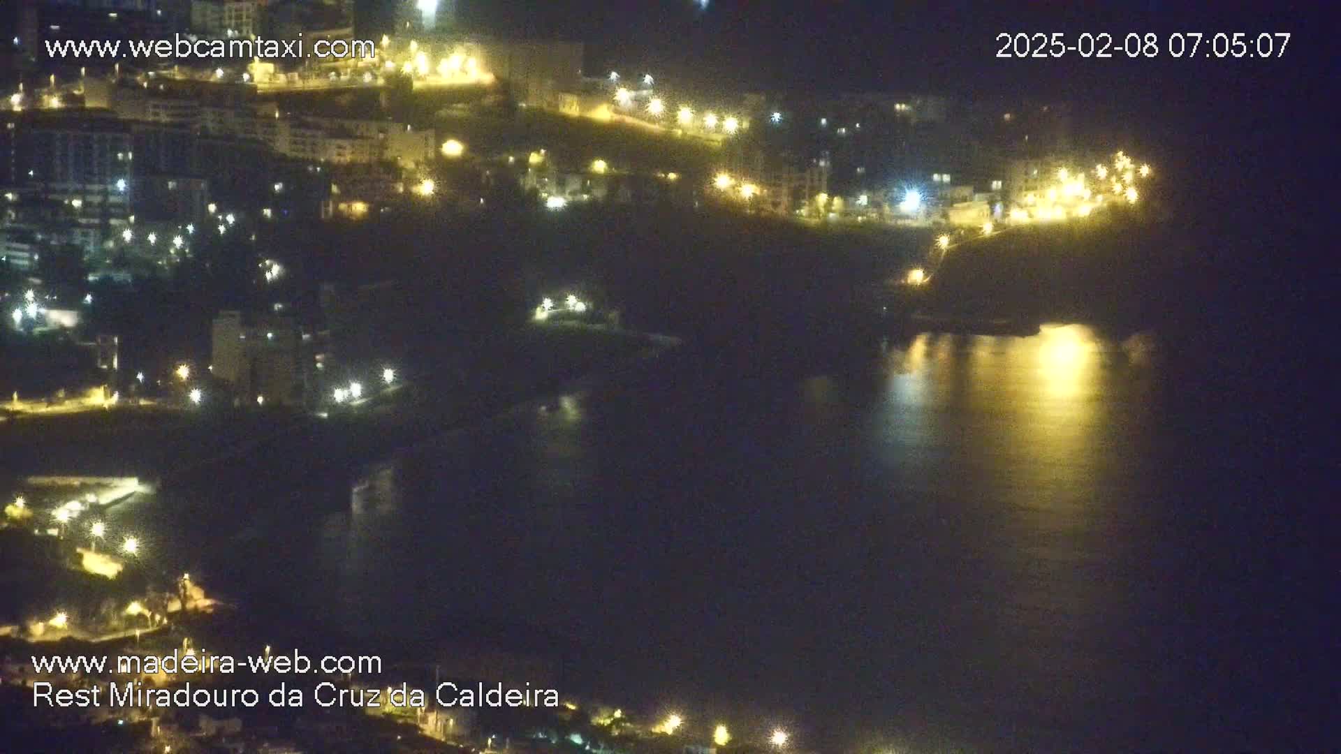 A nighttime, coastal view shows a dark body of water reflecting the lights of buildings on a hillside.