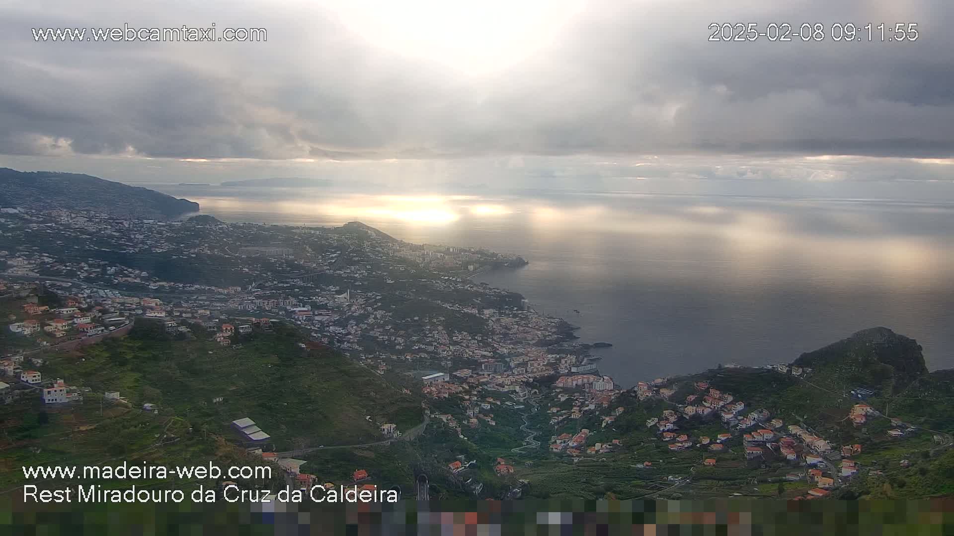 An aerial view shows a coastal town nestled on a hillside overlooking a calm ocean under a mostly cloudy sky with bright sunlight breaking through.