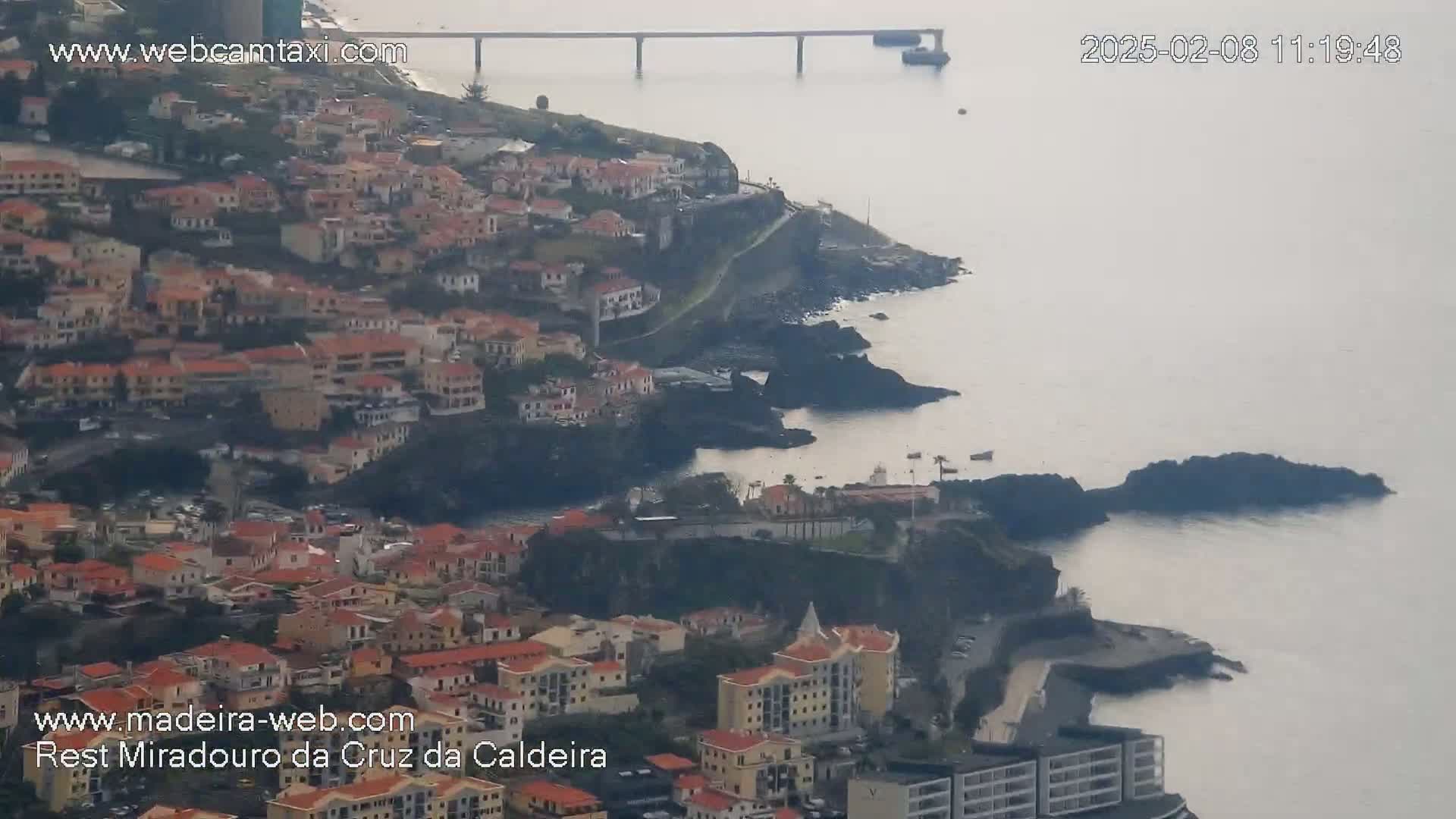 An aerial view shows a coastal town nestled along a rocky coastline, with calm, hazy water extending to a distant bridge under an overcast sky.