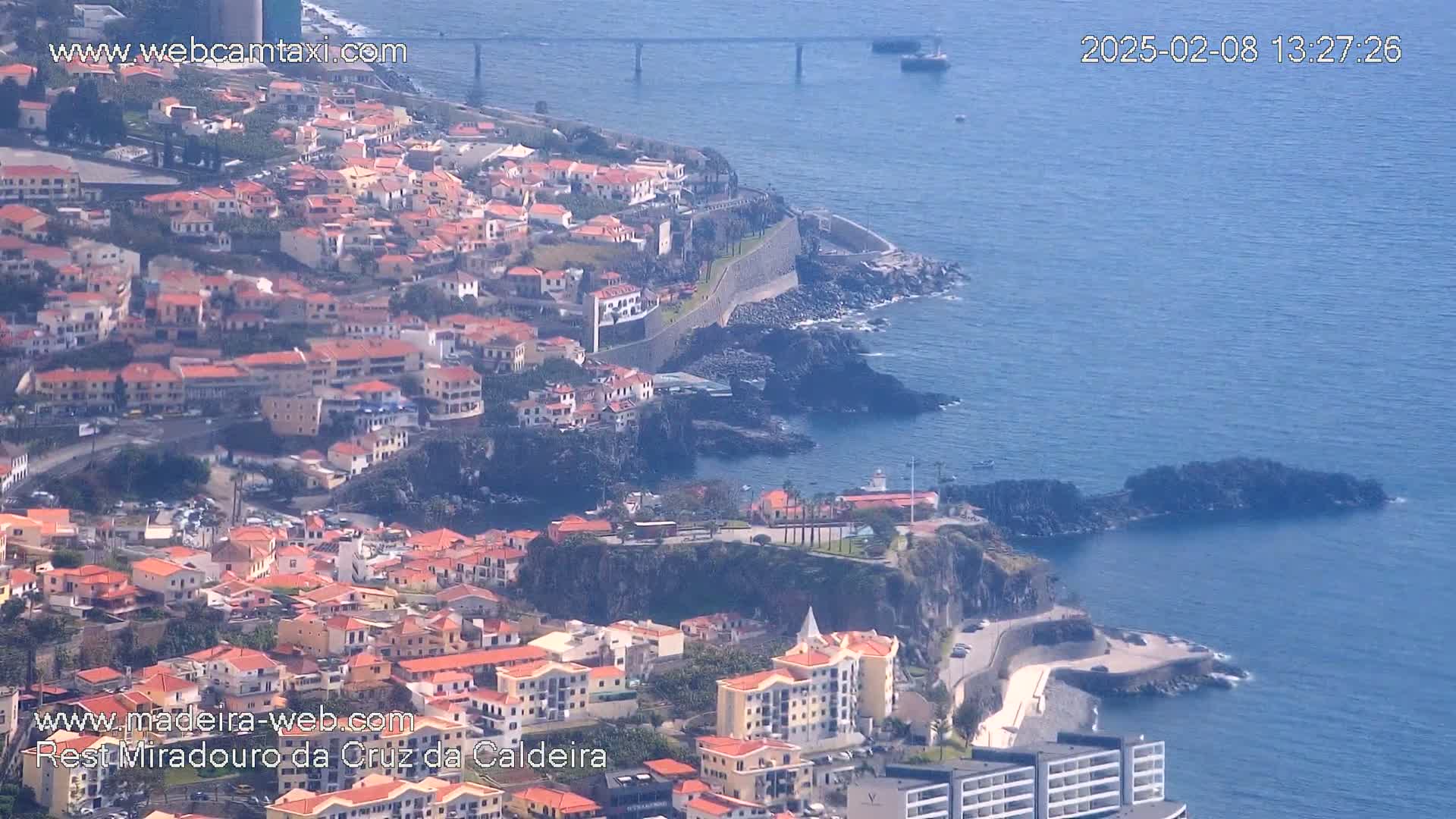 An aerial view shows a coastal town with red-roofed buildings cascading down a cliffside into a calm blue ocean under a clear sky.