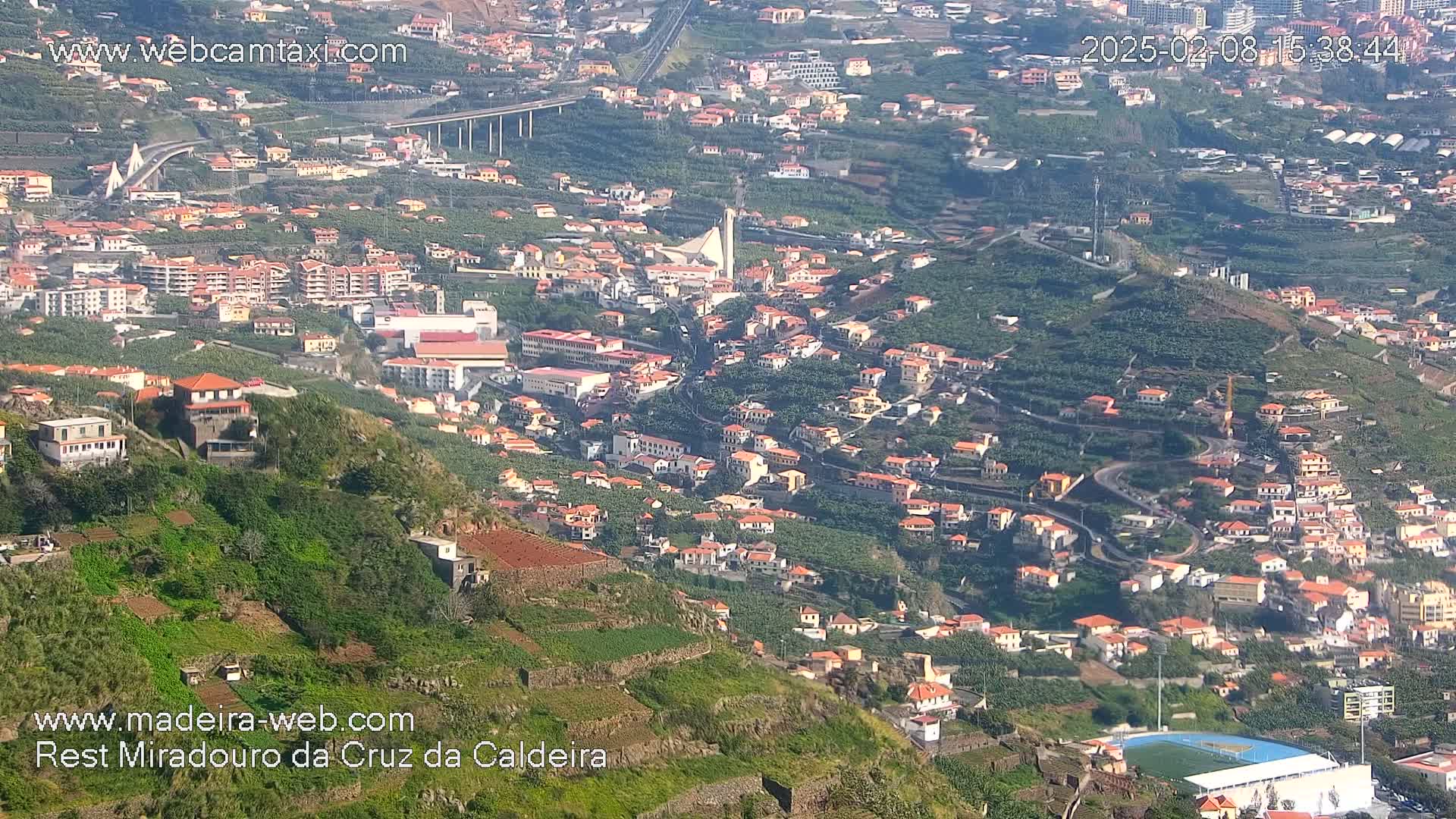 A high-angle view shows a hillside town with many red-roofed buildings nestled among green vegetation on a sunny day.