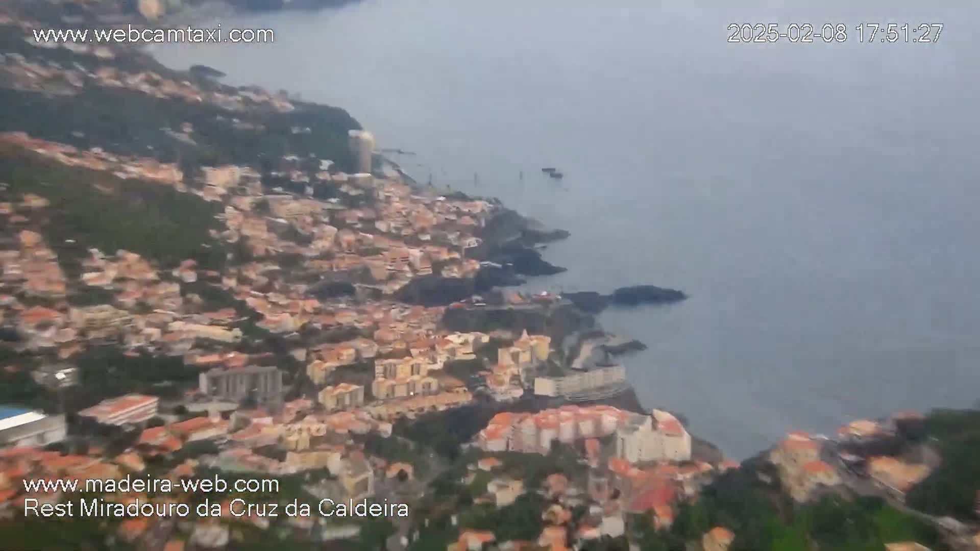A hazy aerial view shows a coastal town nestled on a hillside, meeting a calm ocean.
