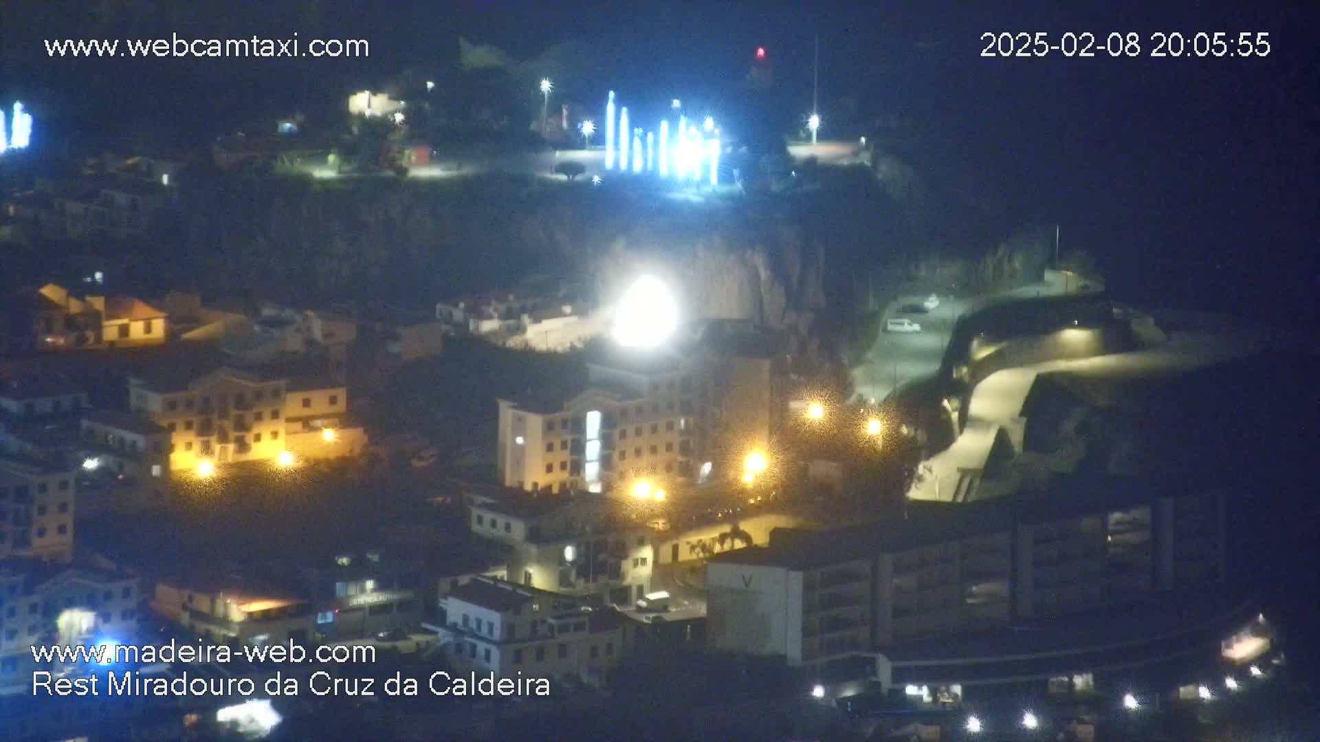 An aerial nighttime view of a town illuminated by streetlights, with a brightly lit structure and several tall, thin lights visible on a hilltop in the background.