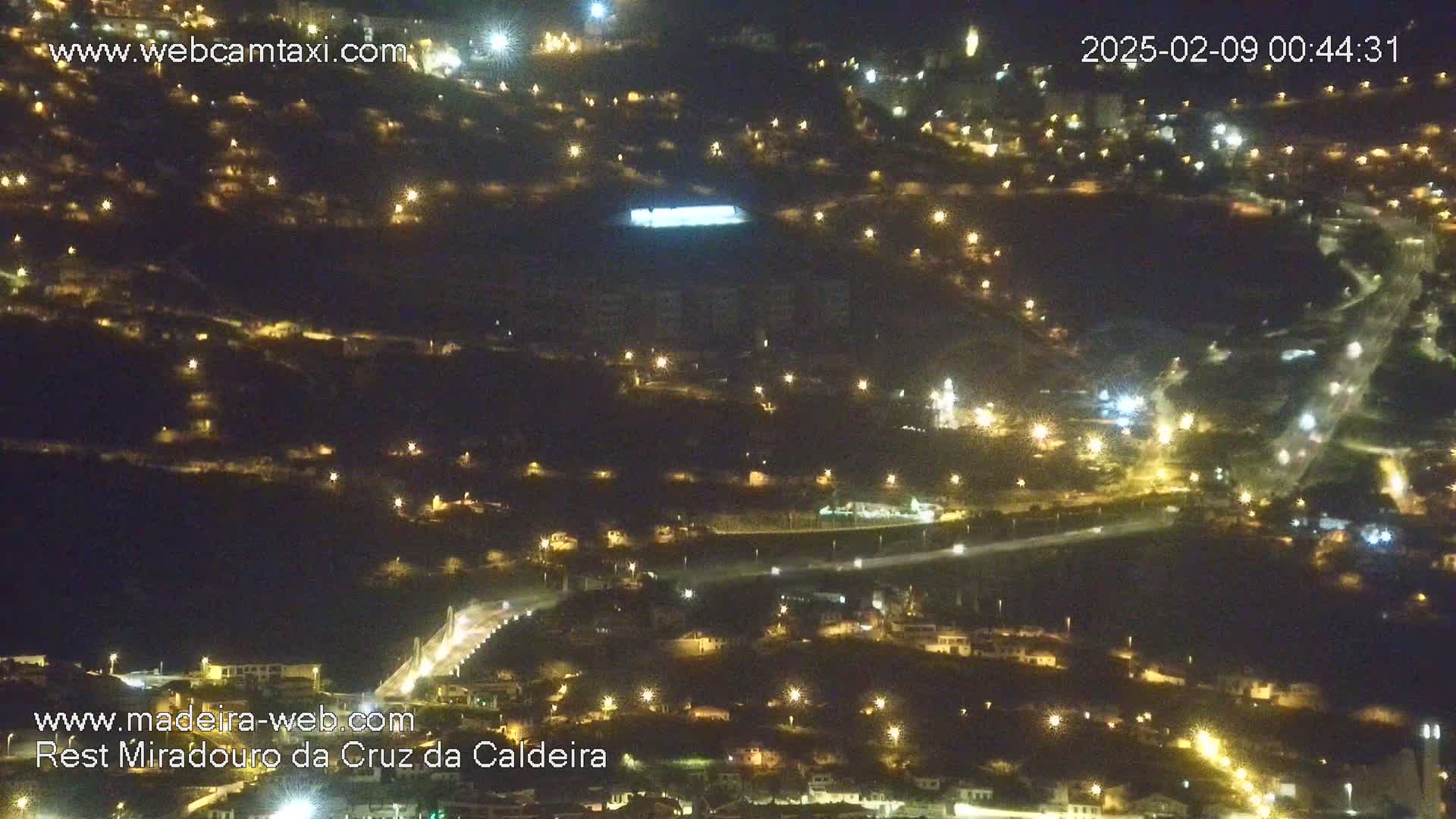 A nighttime aerial view shows a hilly town illuminated by numerous lights, with a road winding through it.