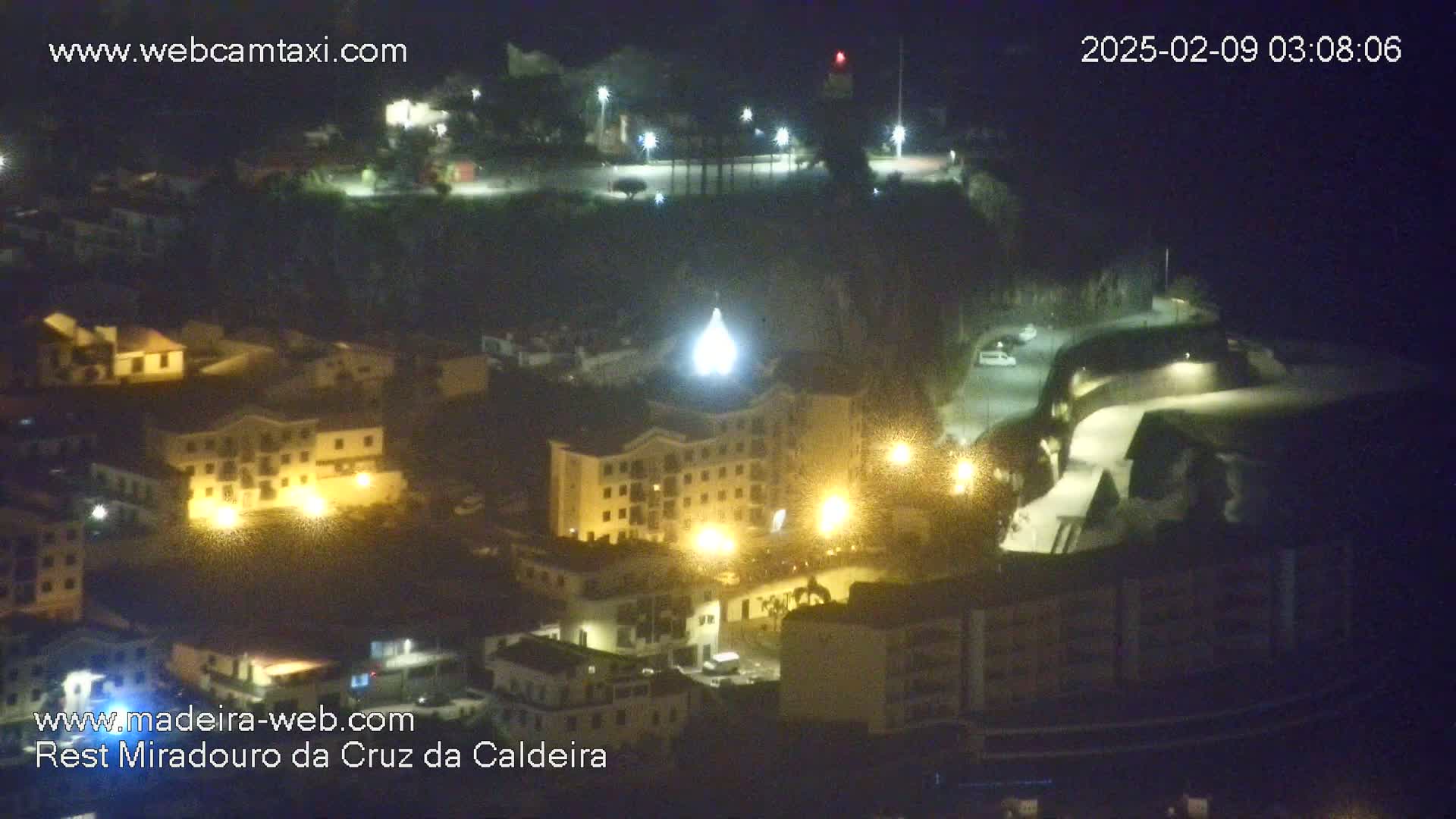 A nighttime, high-angle view of a town illuminated by streetlights, featuring buildings of varying sizes and a prominent structure with a pointed top.