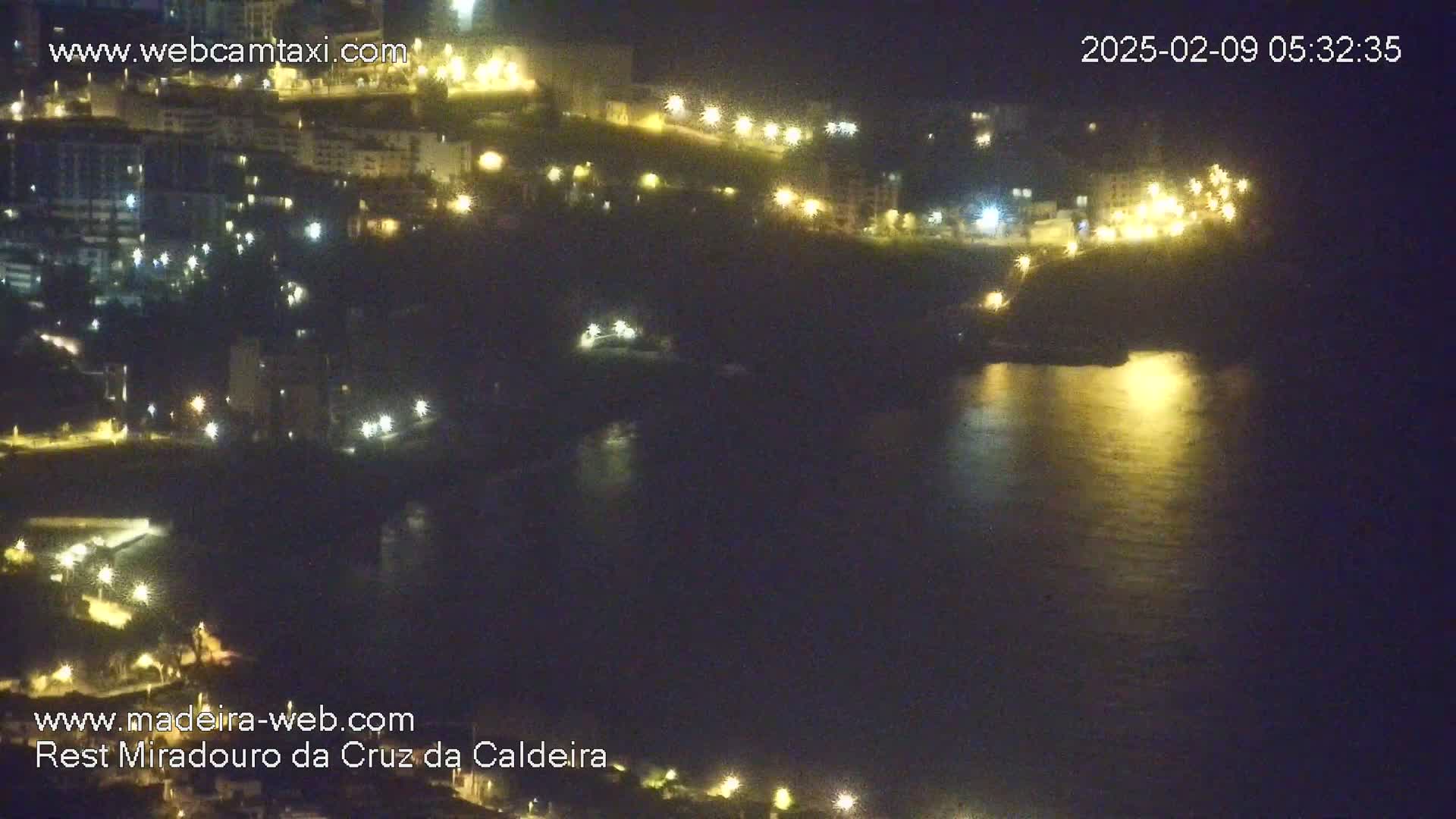 A nighttime view of a coastline with buildings illuminated by lights, and a dark body of water reflecting the city lights.